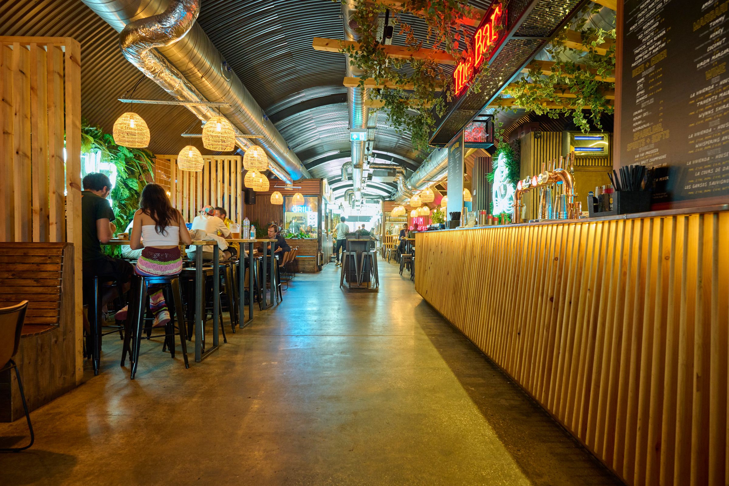 An industrial-chic food hall with a corrugated ceiling, exposed ducts, wooden paneling, people dining, and a long bar.