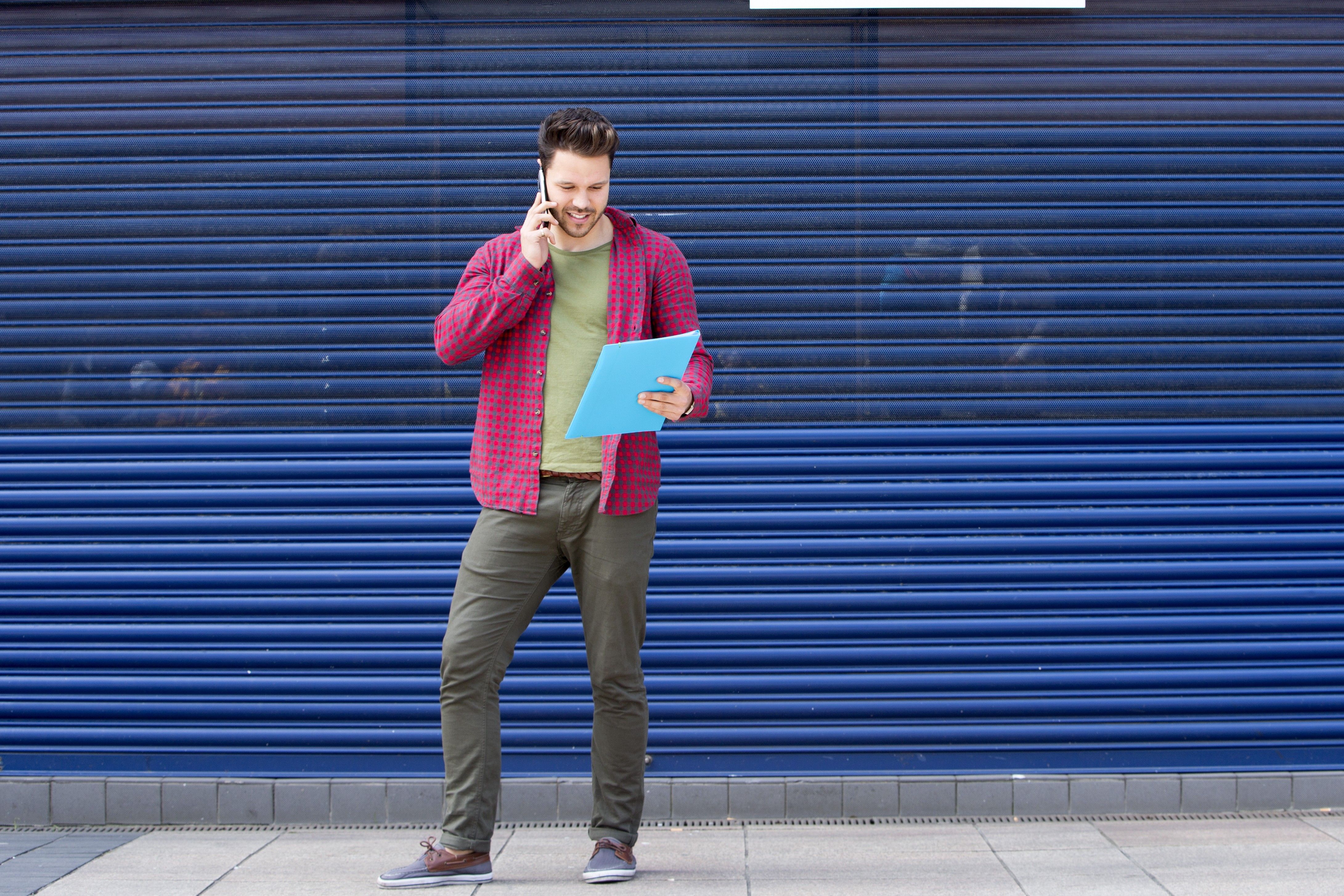 A man talks on his phone while holding a blue folder, standing against a blue corrugated wall.