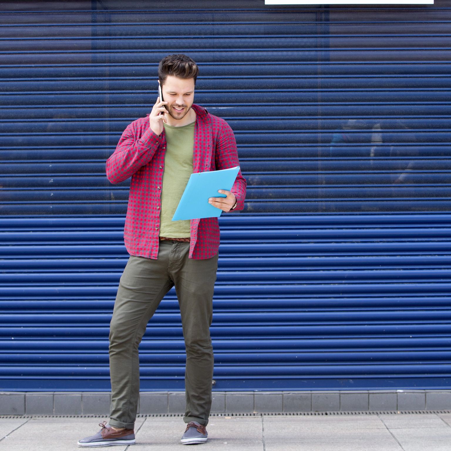 A man talks on his phone while holding a blue folder, standing against a blue corrugated wall.
