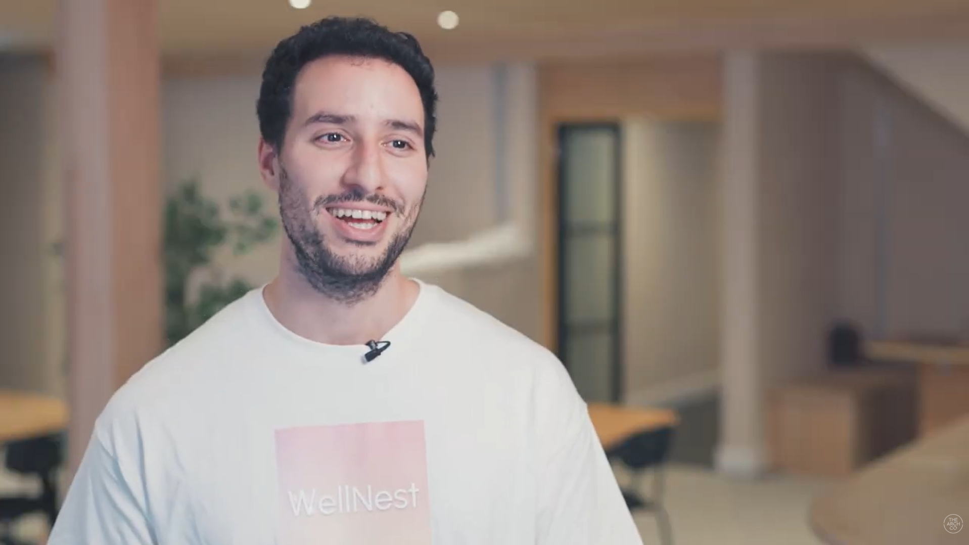 A smiling man in a white "WellNest" t-shirt with a lavalier microphone in a modern office.