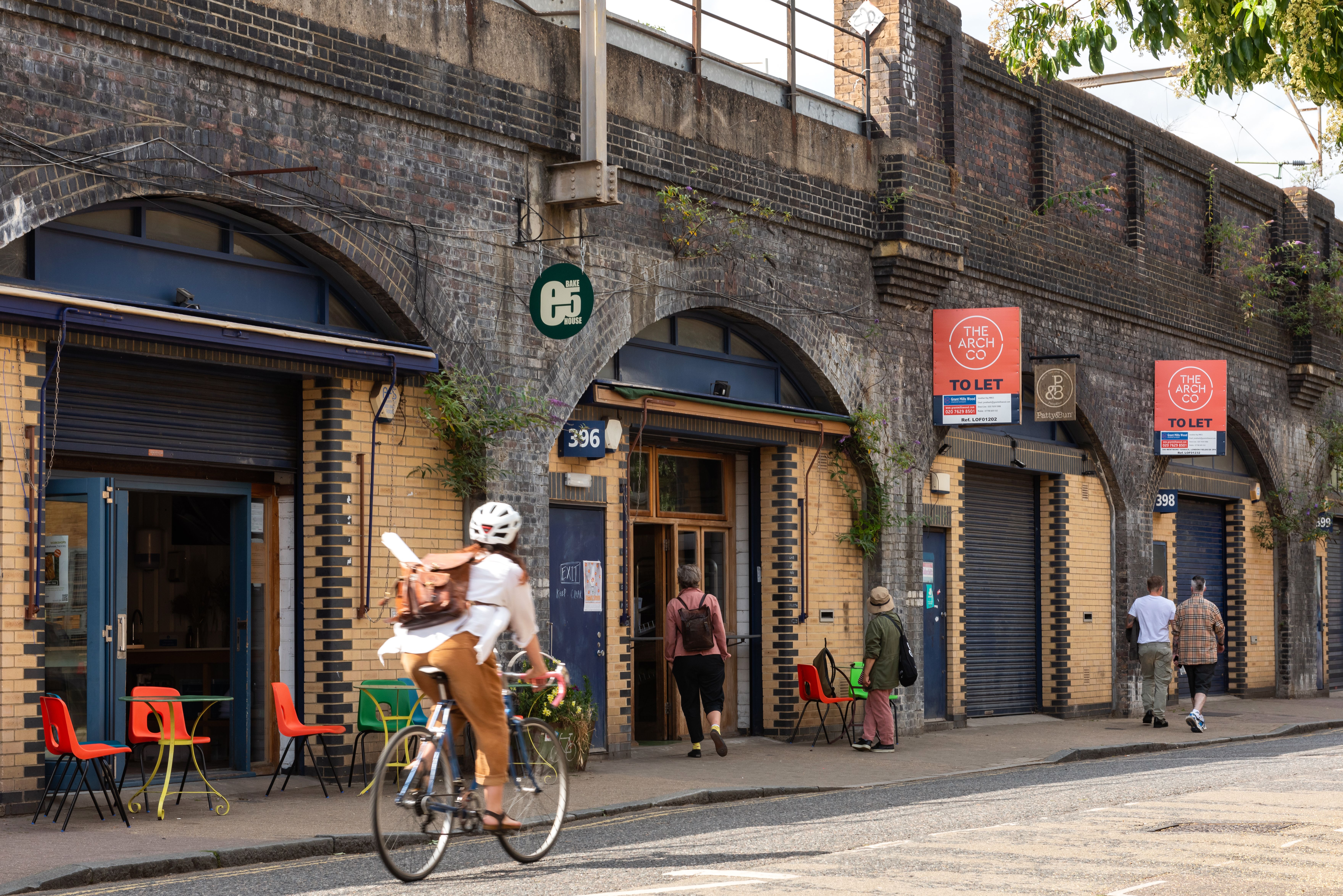 A street scene with people walking and cycling past businesses and 'To Let' units in historic brick railway arches.
