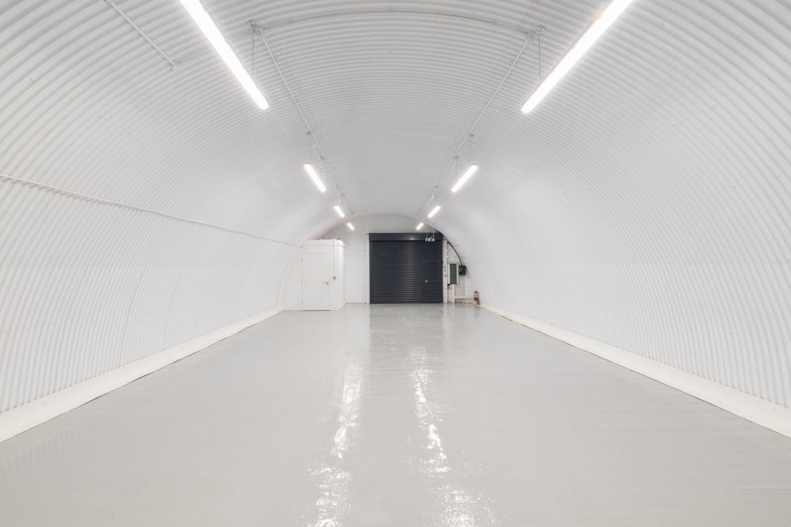 A brightly lit, long, white corrugated arched tunnel with a light gray floor and a dark door at the end.
