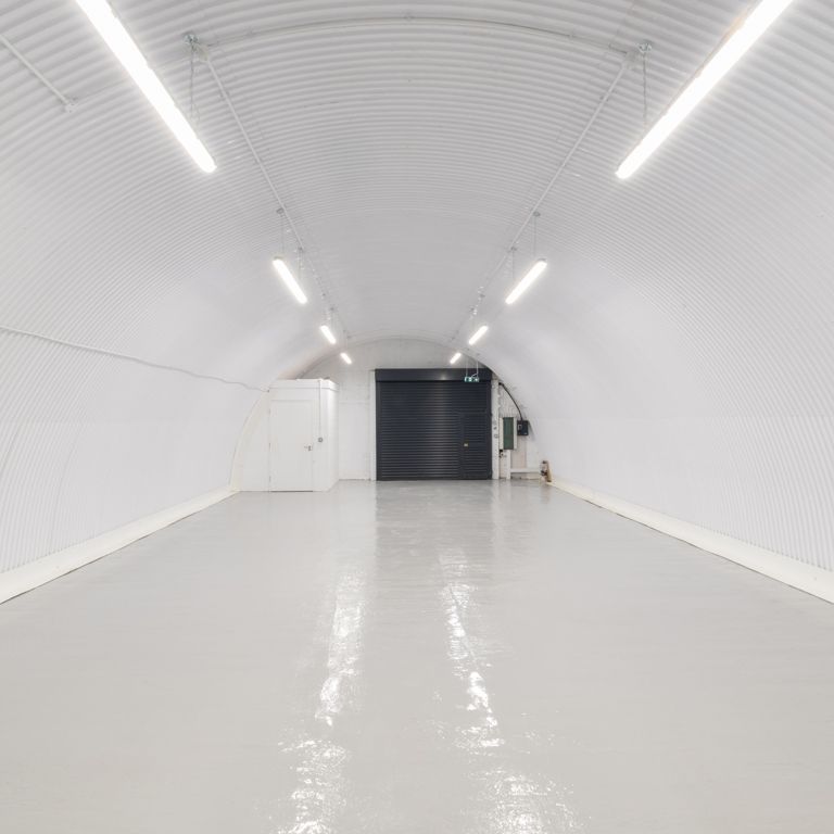 A brightly lit, long, white corrugated arched tunnel with a light gray floor and a dark door at the end.