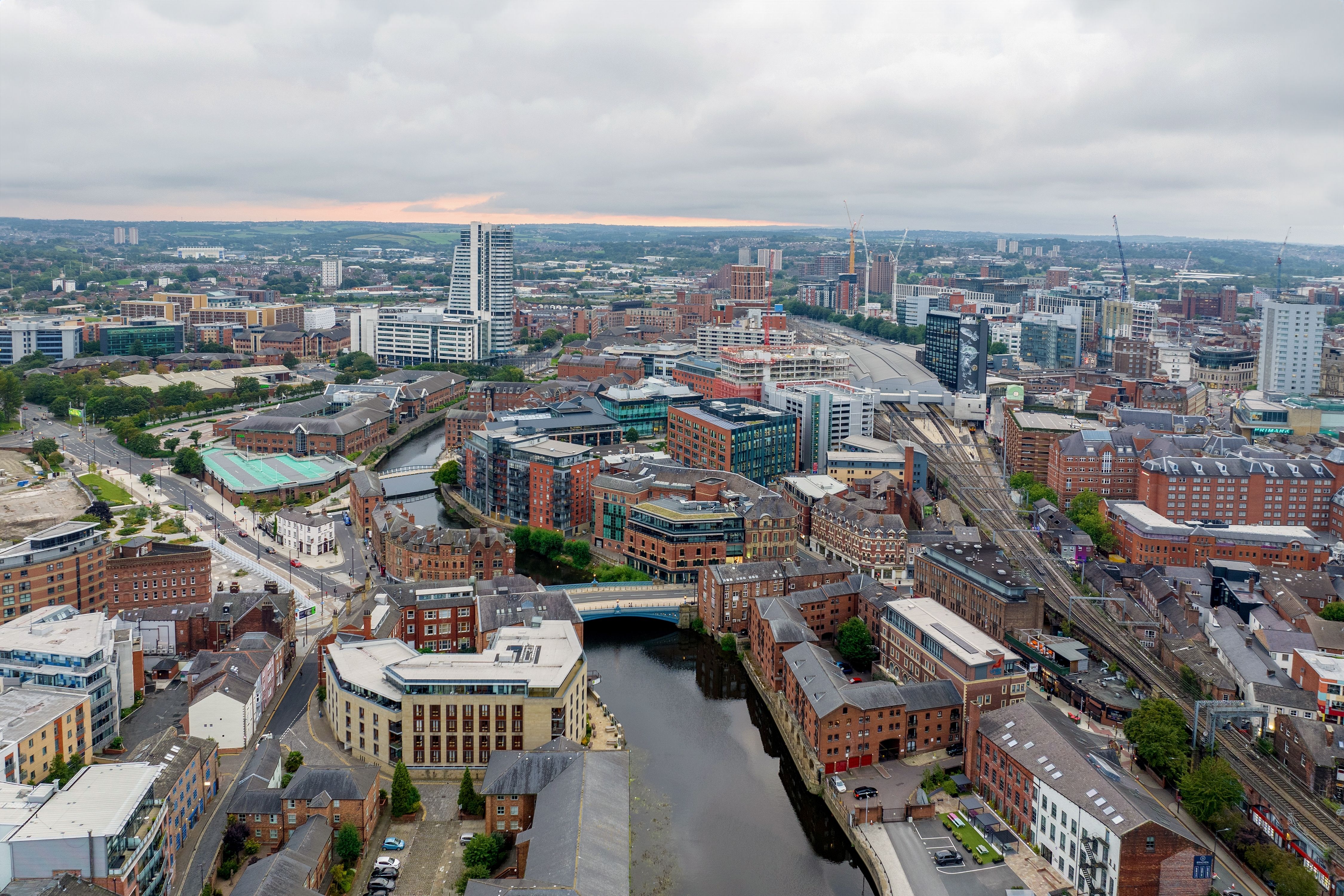 Aerial view of a city with a winding river, modern high-rises, historic brick buildings, and train tracks under a cloudy sky.