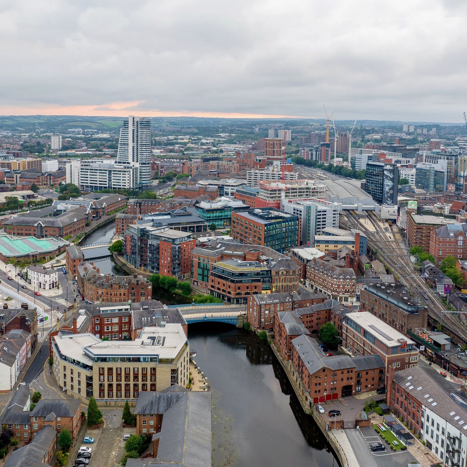 Aerial view of a city with a winding river, modern high-rises, historic brick buildings, and train tracks under a cloudy sky.