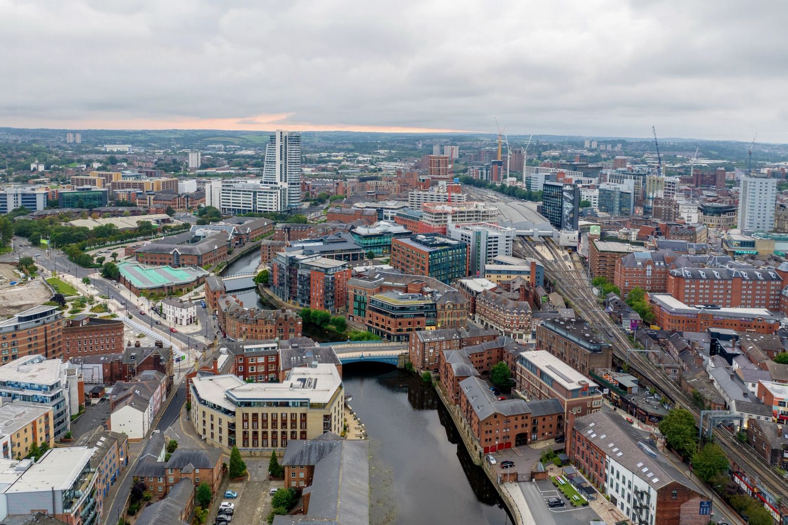 Aerial view of a city with a winding river, modern high-rises, historic brick buildings, and train tracks under a cloudy sky.