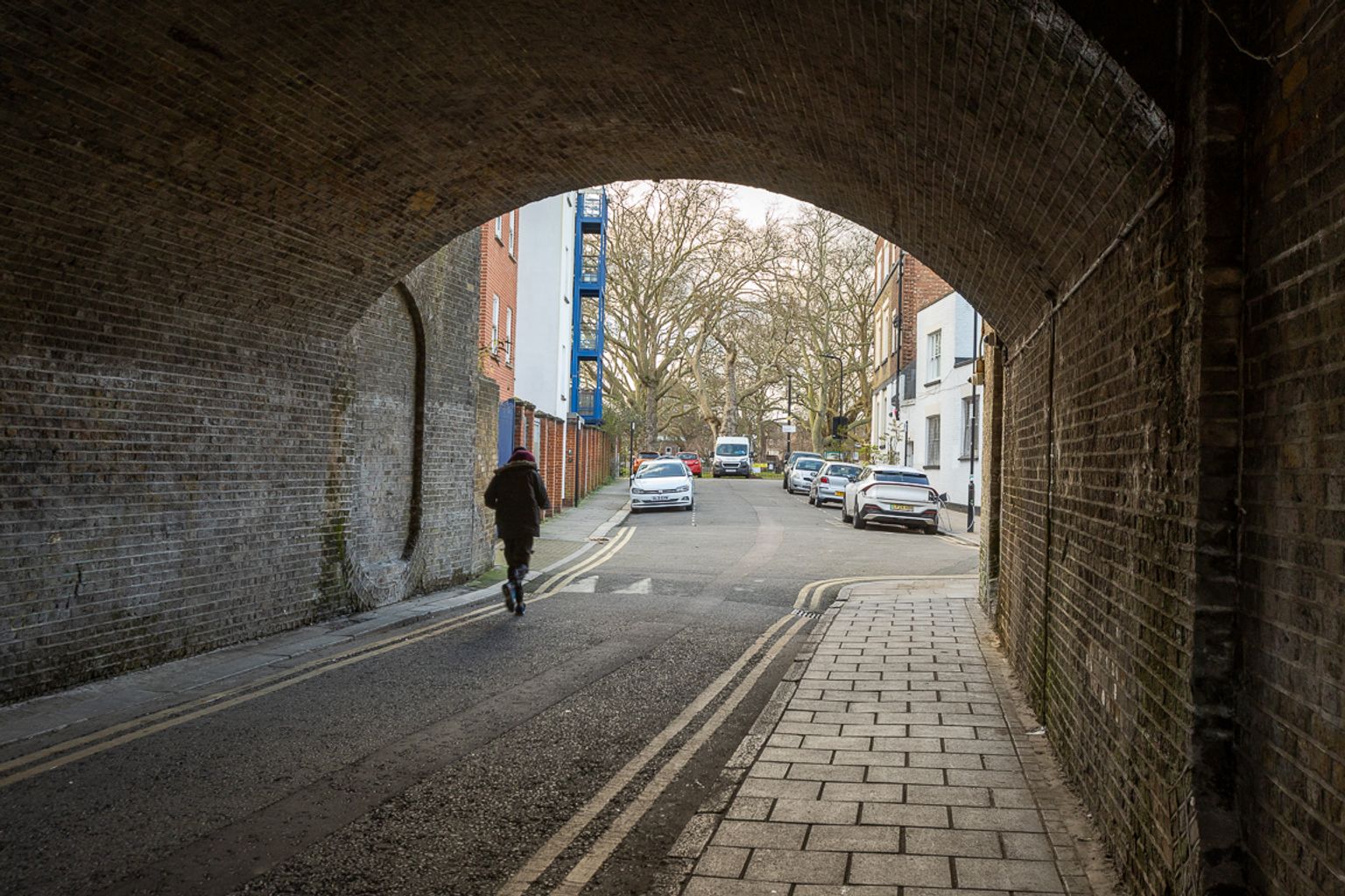 A person walks through a brick archway onto a street with parked cars and buildings.