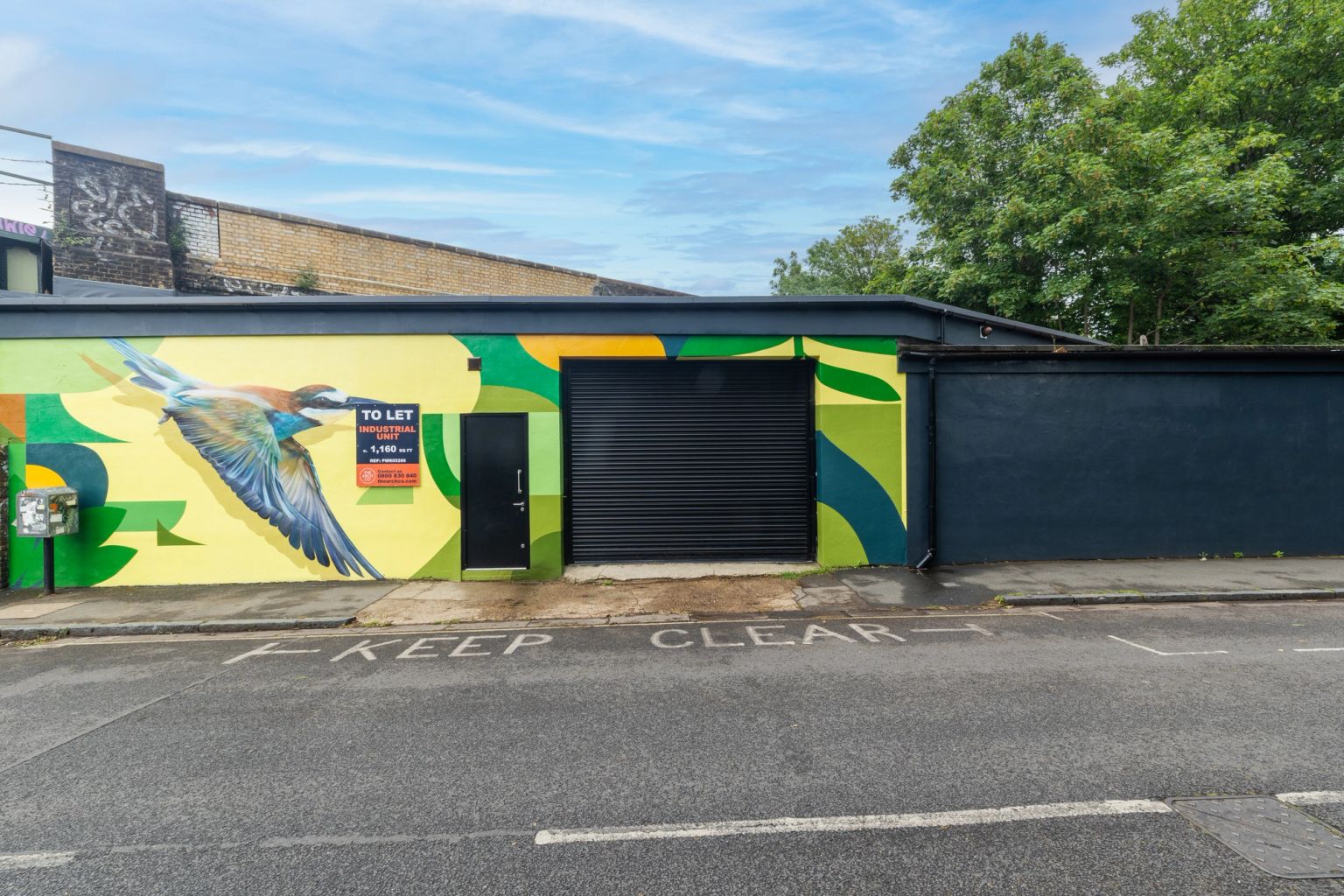 A building with a colorful bird mural, a "To Let" sign, and a black roller door.