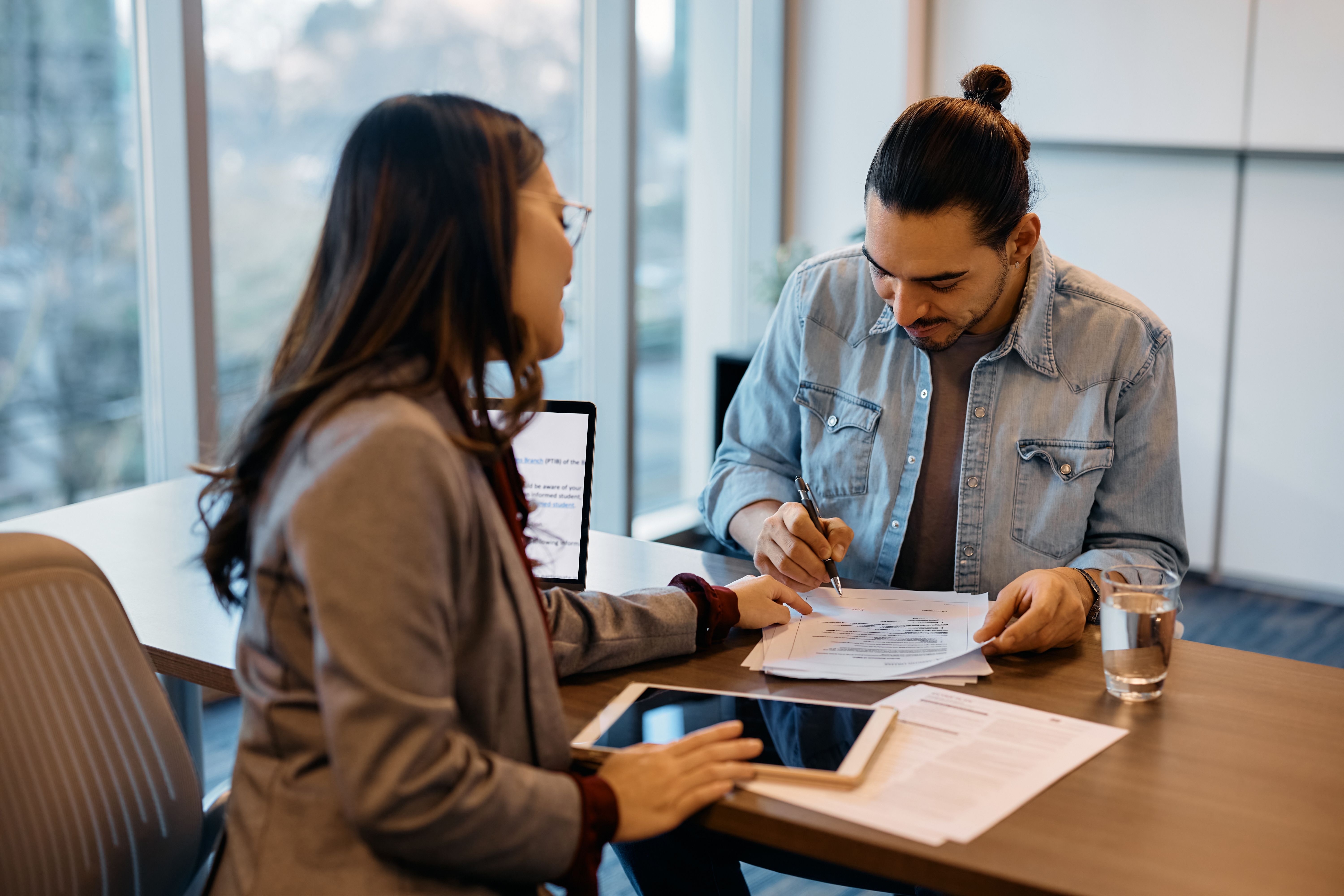 A man with a bun signs a document at a desk while a woman in glasses looks on.