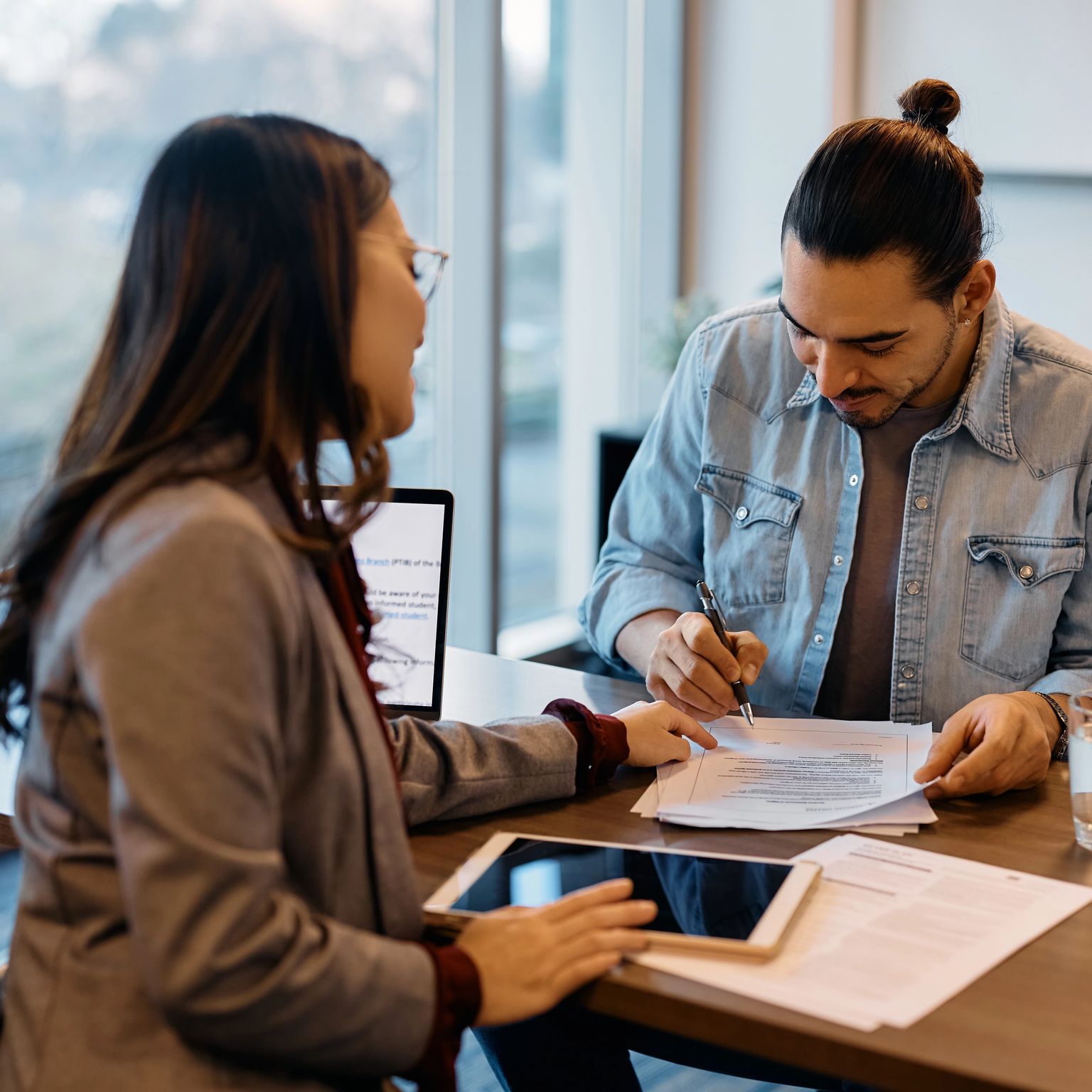 A man with a bun signs a document at a desk while a woman in glasses looks on.