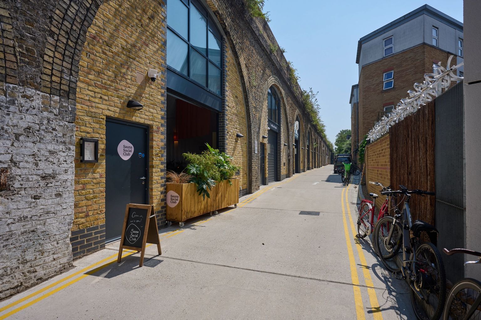 A bright street lined with businesses in brick railway arches, including the Bermondsey Social Club, and bicycles parked along the right.