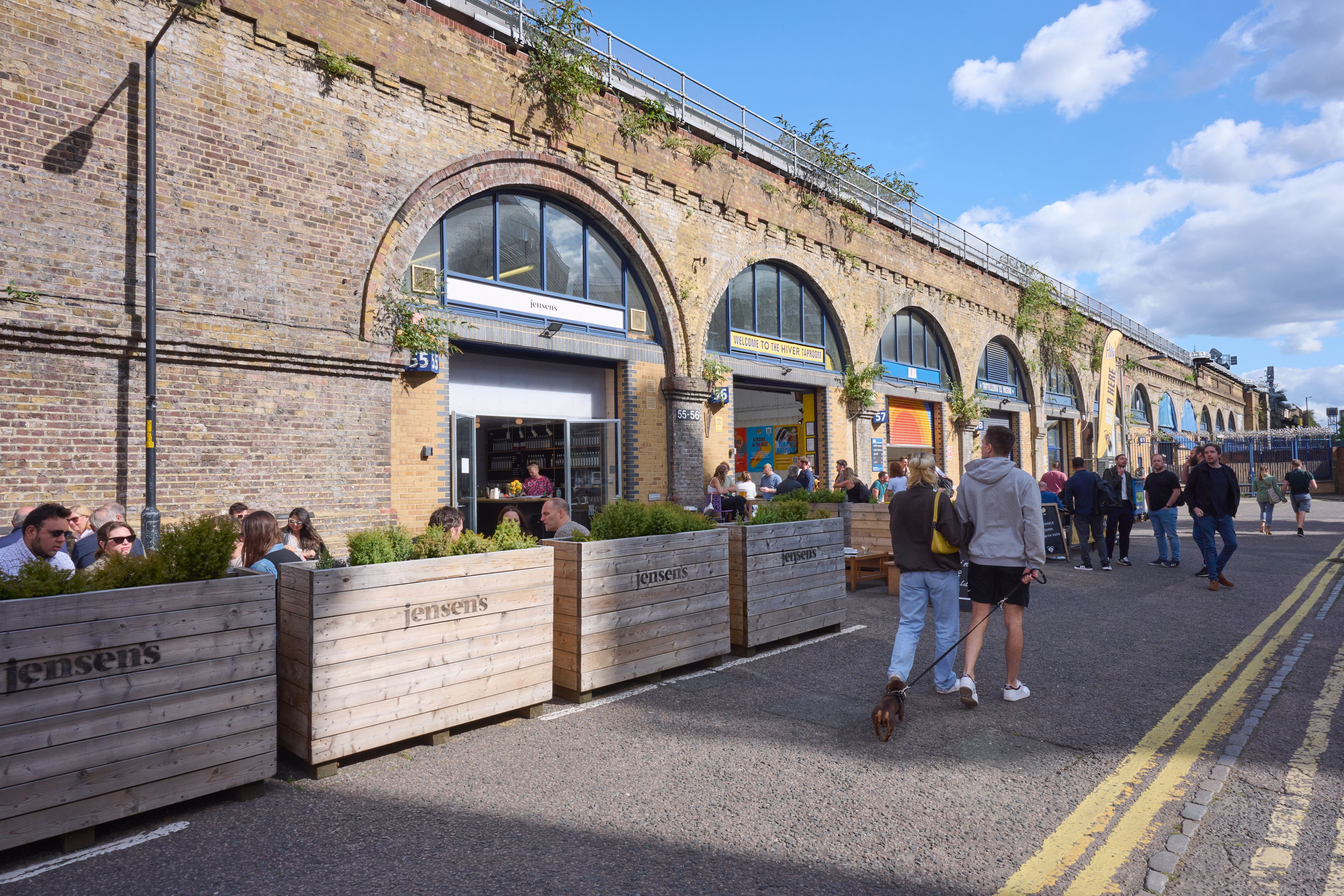 People dine and walk along a street with businesses set in brick railway arches under a blue sky.
