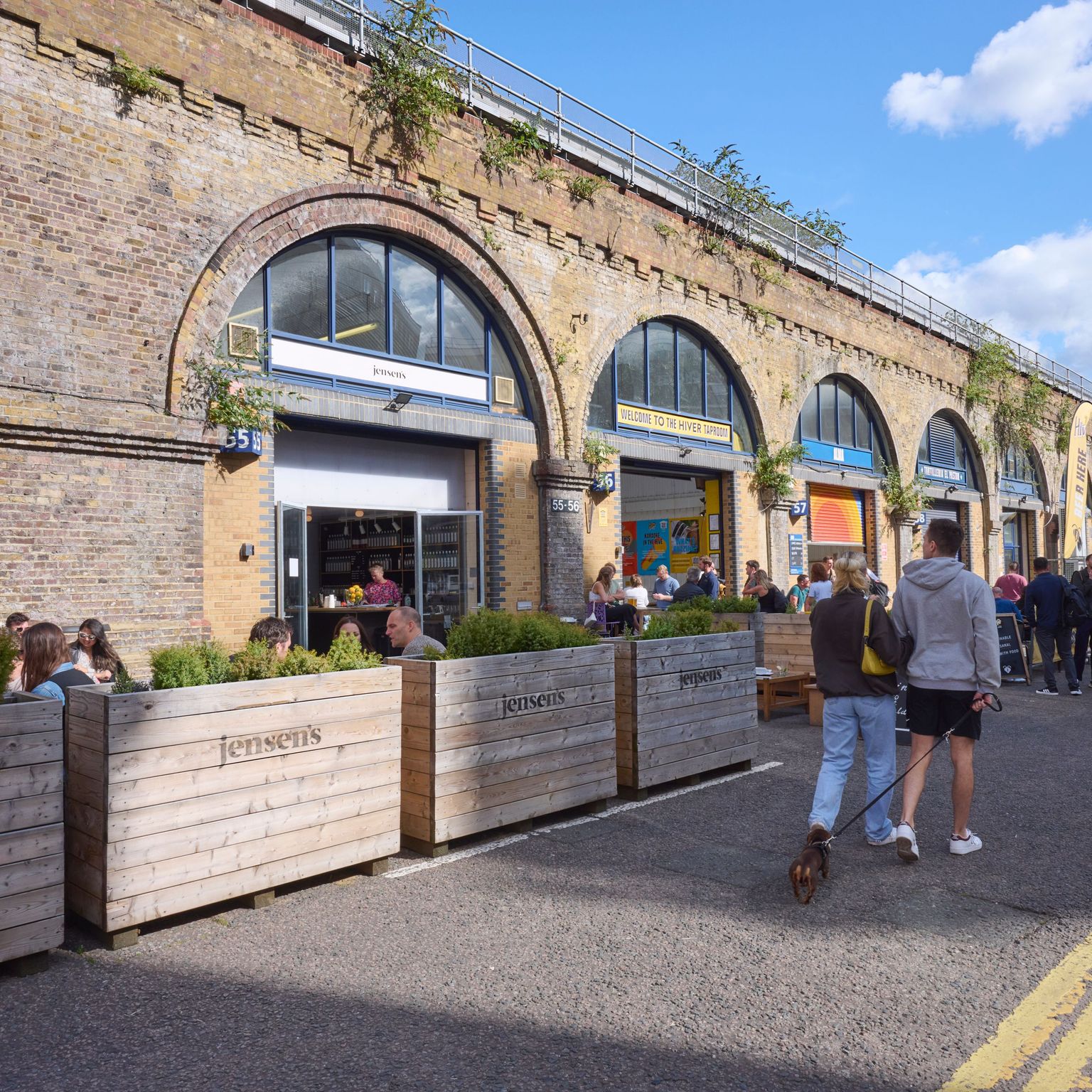 People dine and walk along a street with businesses set in brick railway arches under a blue sky.