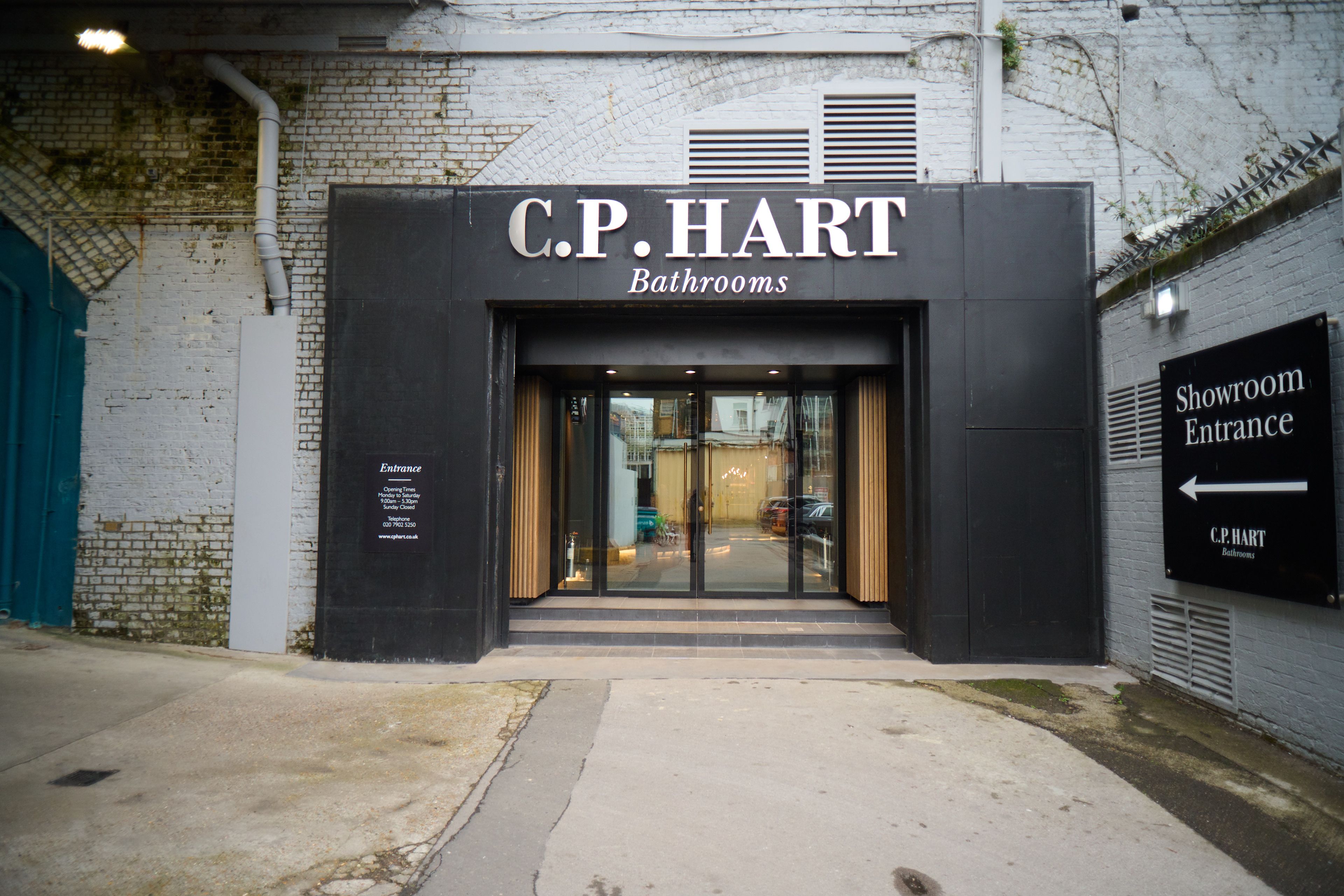 Modern black facade and glass doors of the C.P. HART Bathrooms showroom entrance, with a "Showroom Entrance" sign on a light brick wall.