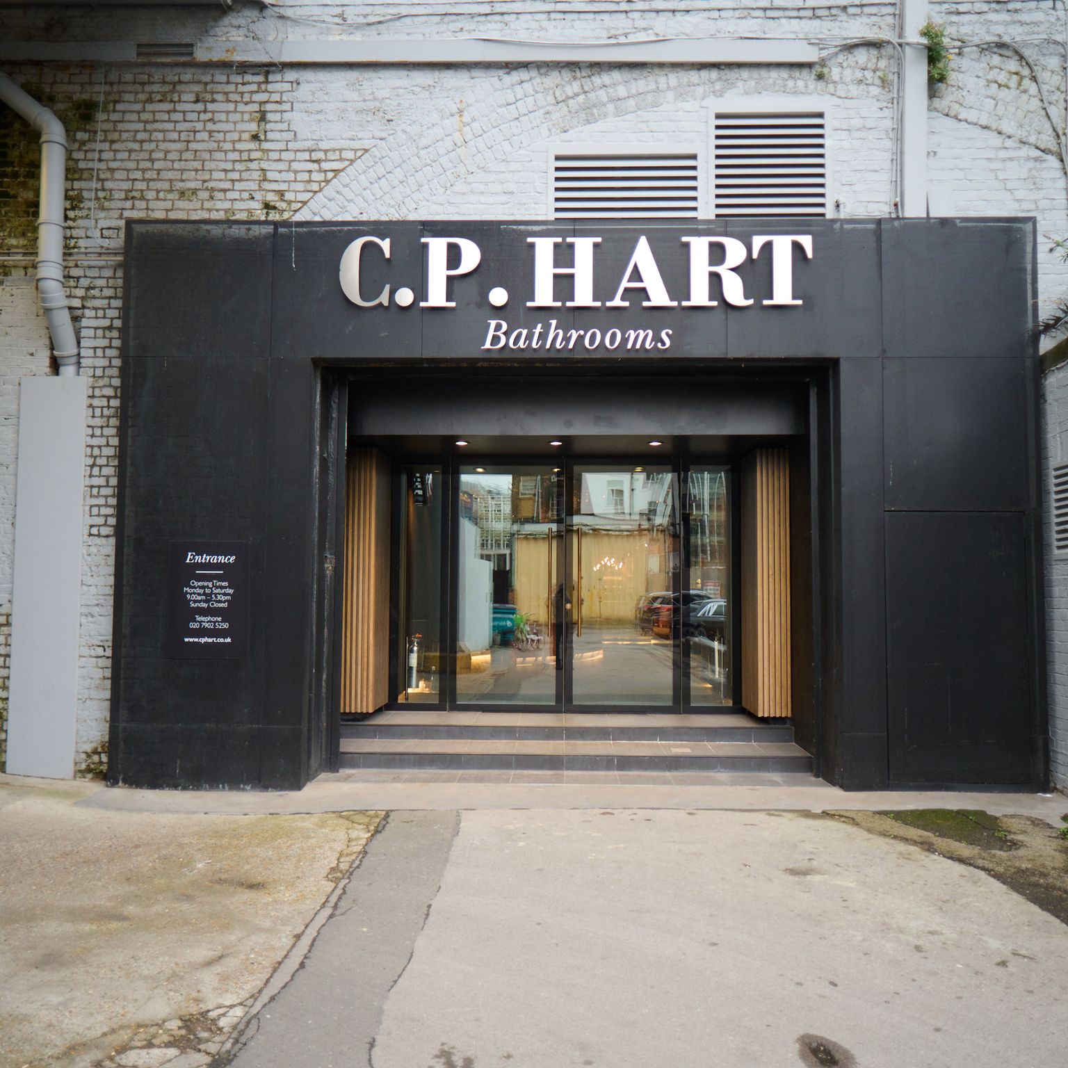 Modern black facade and glass doors of the C.P. HART Bathrooms showroom entrance, with a "Showroom Entrance" sign on a light brick wall.