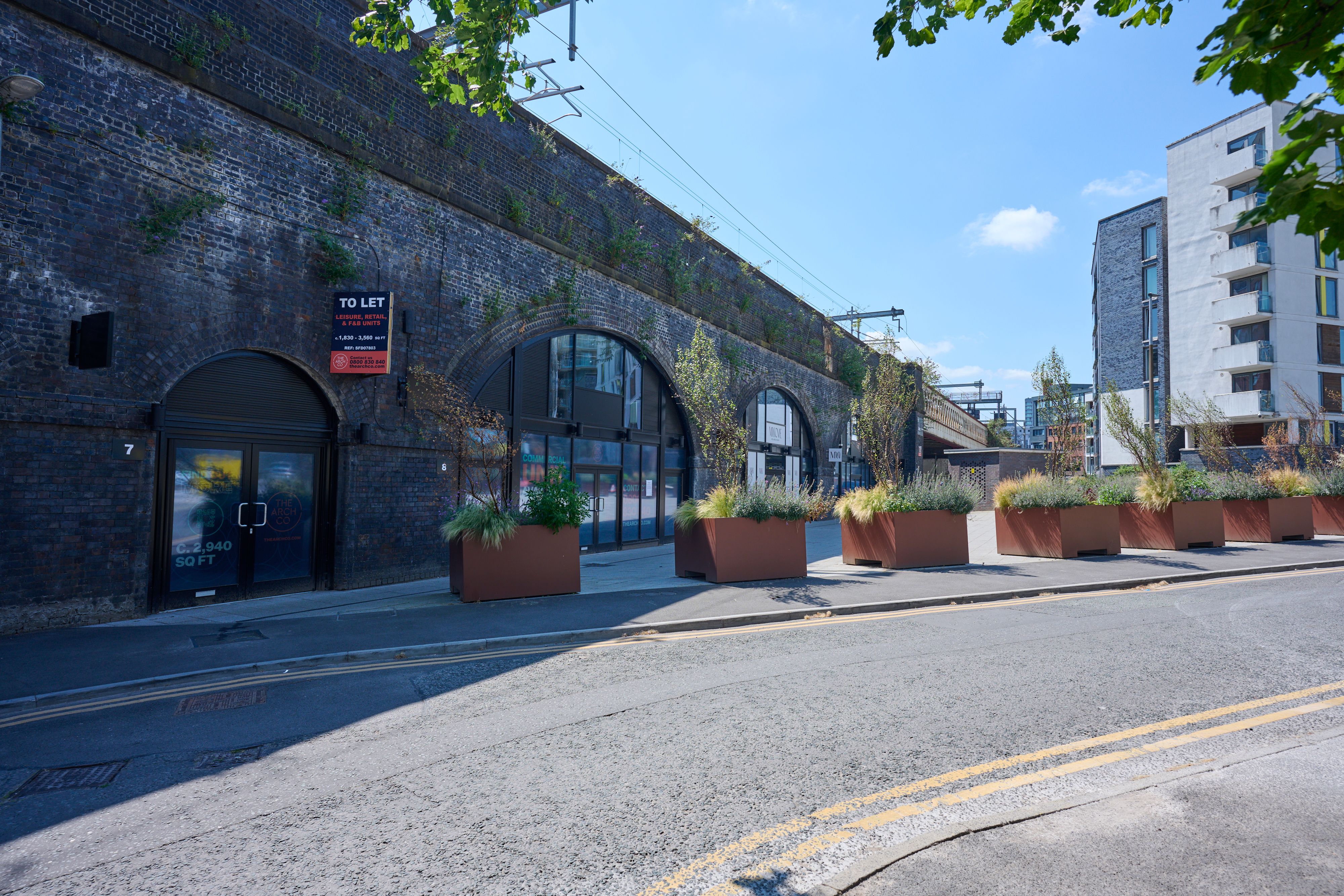 Dark brick railway arches with storefronts and a "TO LET" sign, fronted by a row of planters, alongside a modern building.