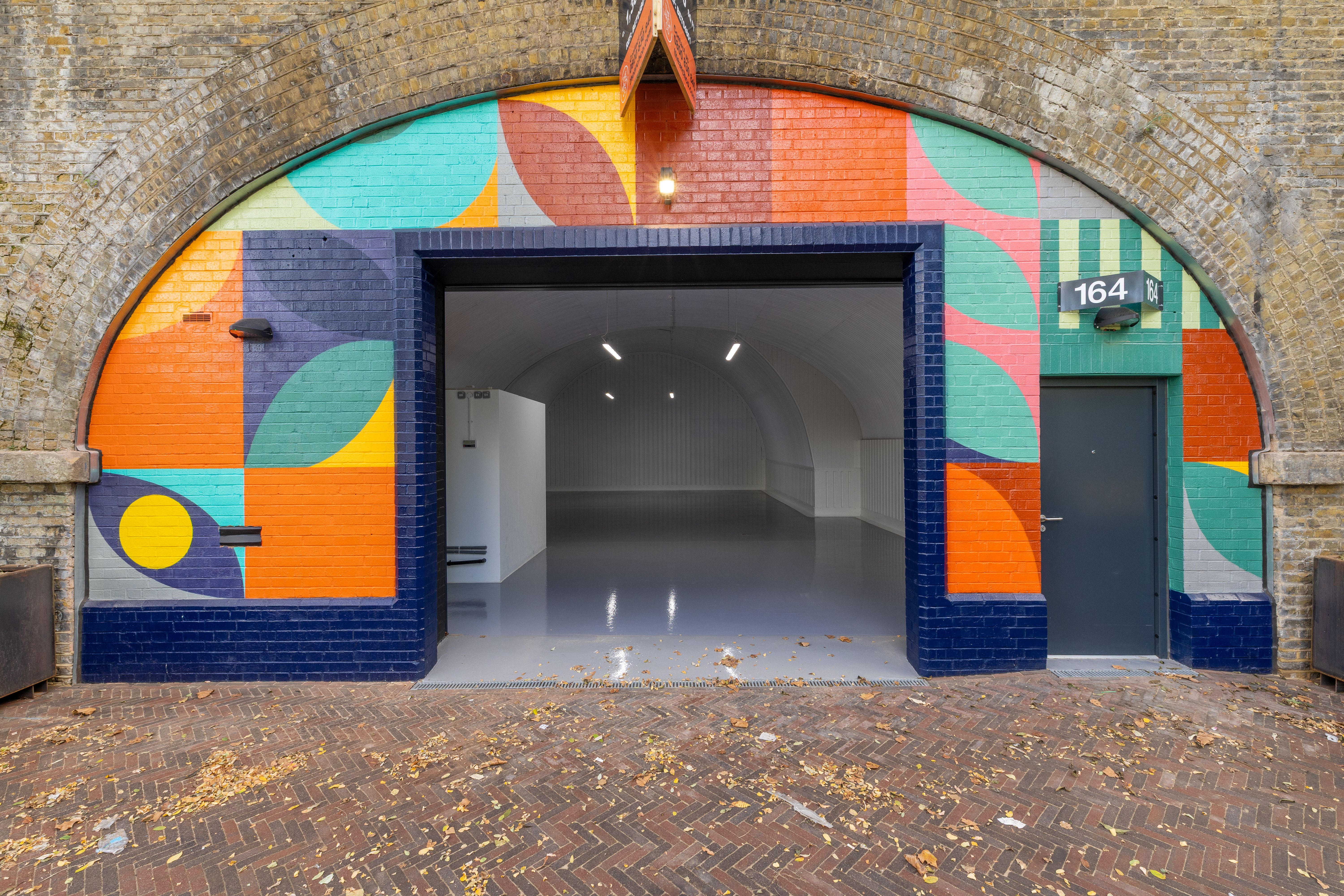 A colorful geometric mural decorates a brick archway entrance, featuring a large opening to a modern white interior and a smaller door labeled 164 B.