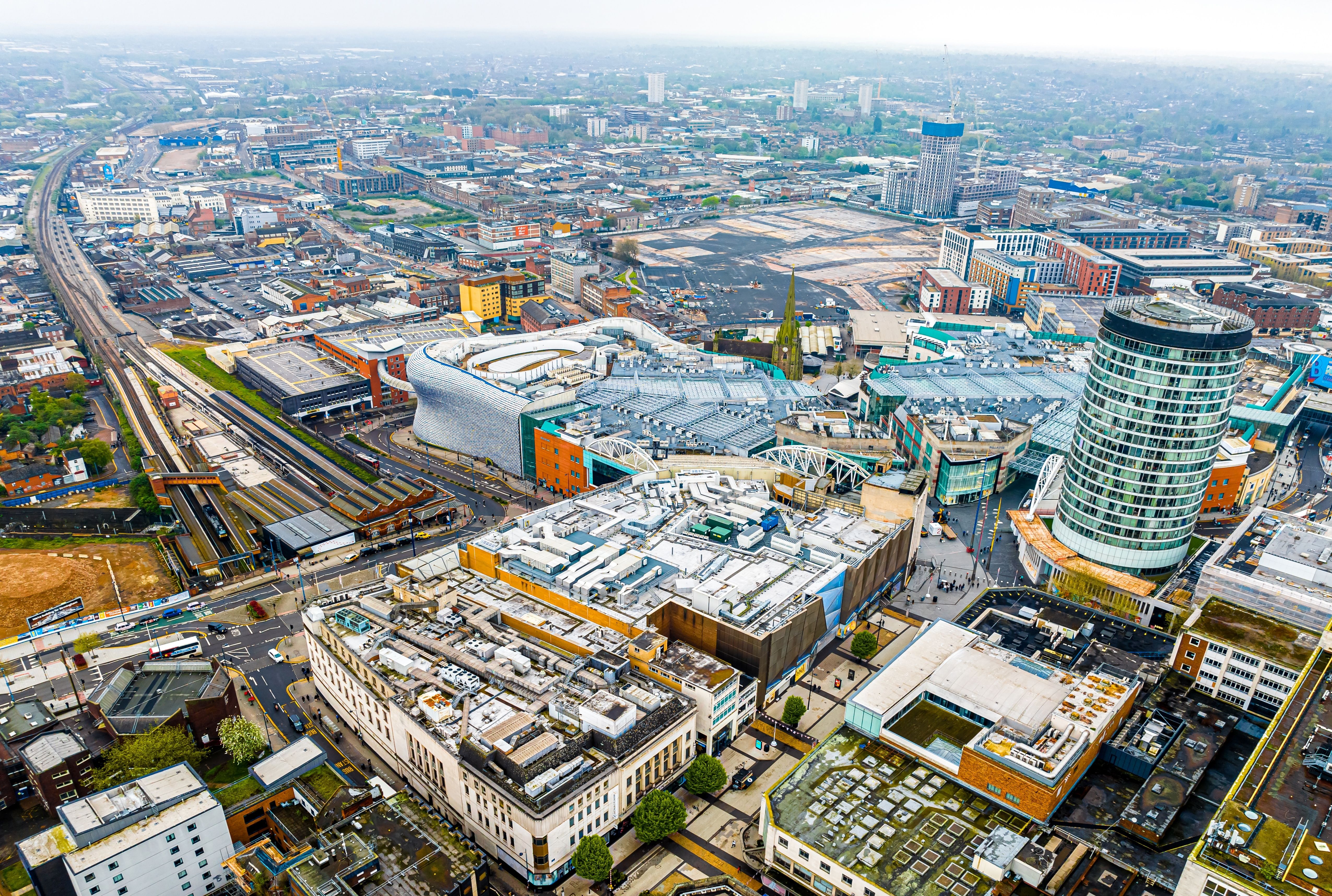 Aerial view of Birmingham city center featuring the distinct Selfridges building, the cylindrical Rotunda, a major train station, and dense urban development.