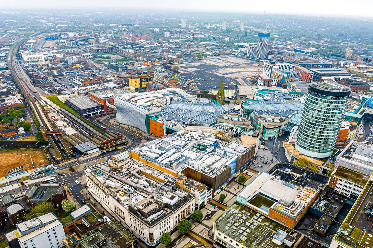 Aerial view of Birmingham city center featuring the distinct Selfridges building, the cylindrical Rotunda, a major train station, and dense urban development.