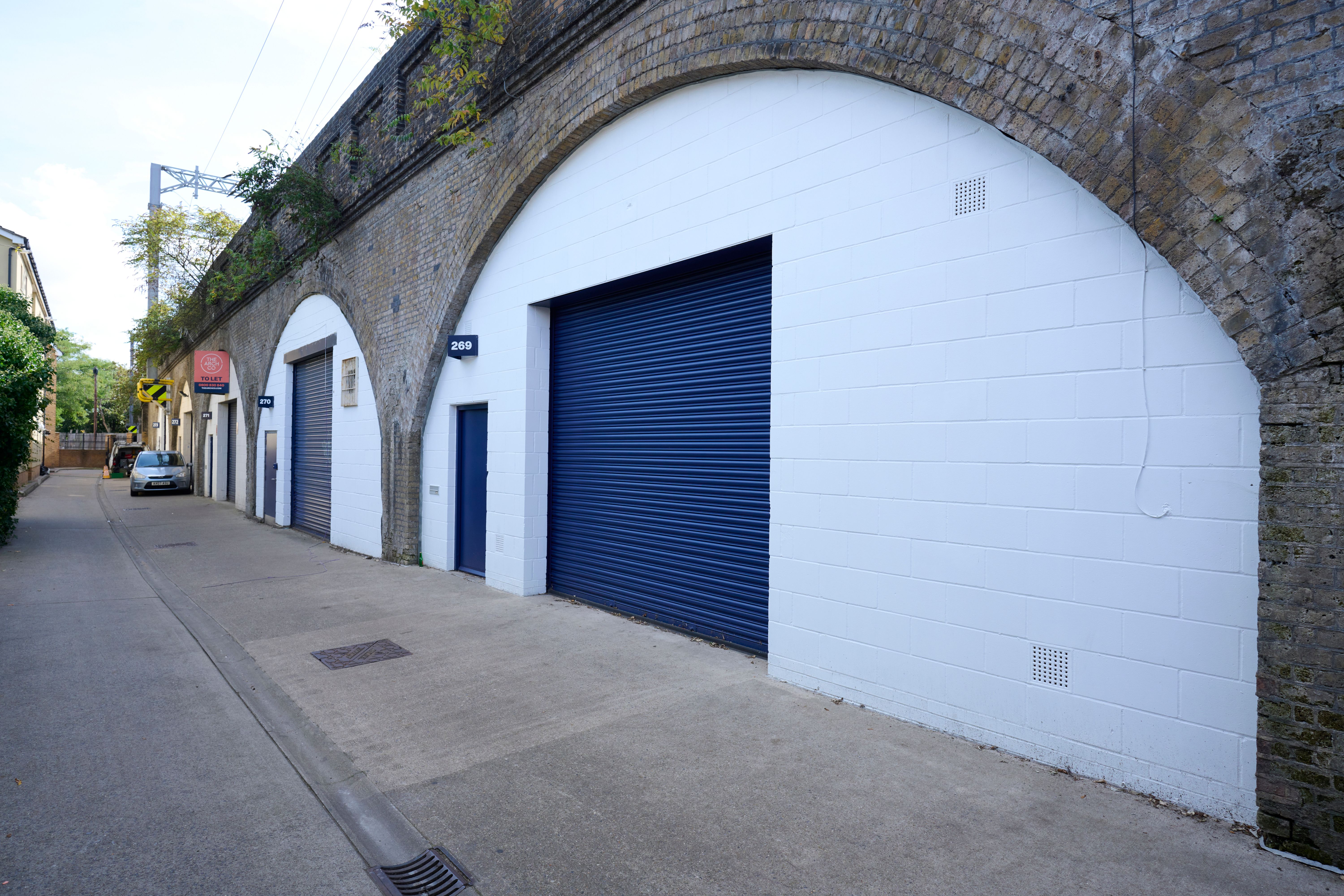 A narrow street alongside brick railway arches converted into white units with dark blue roller shutters.