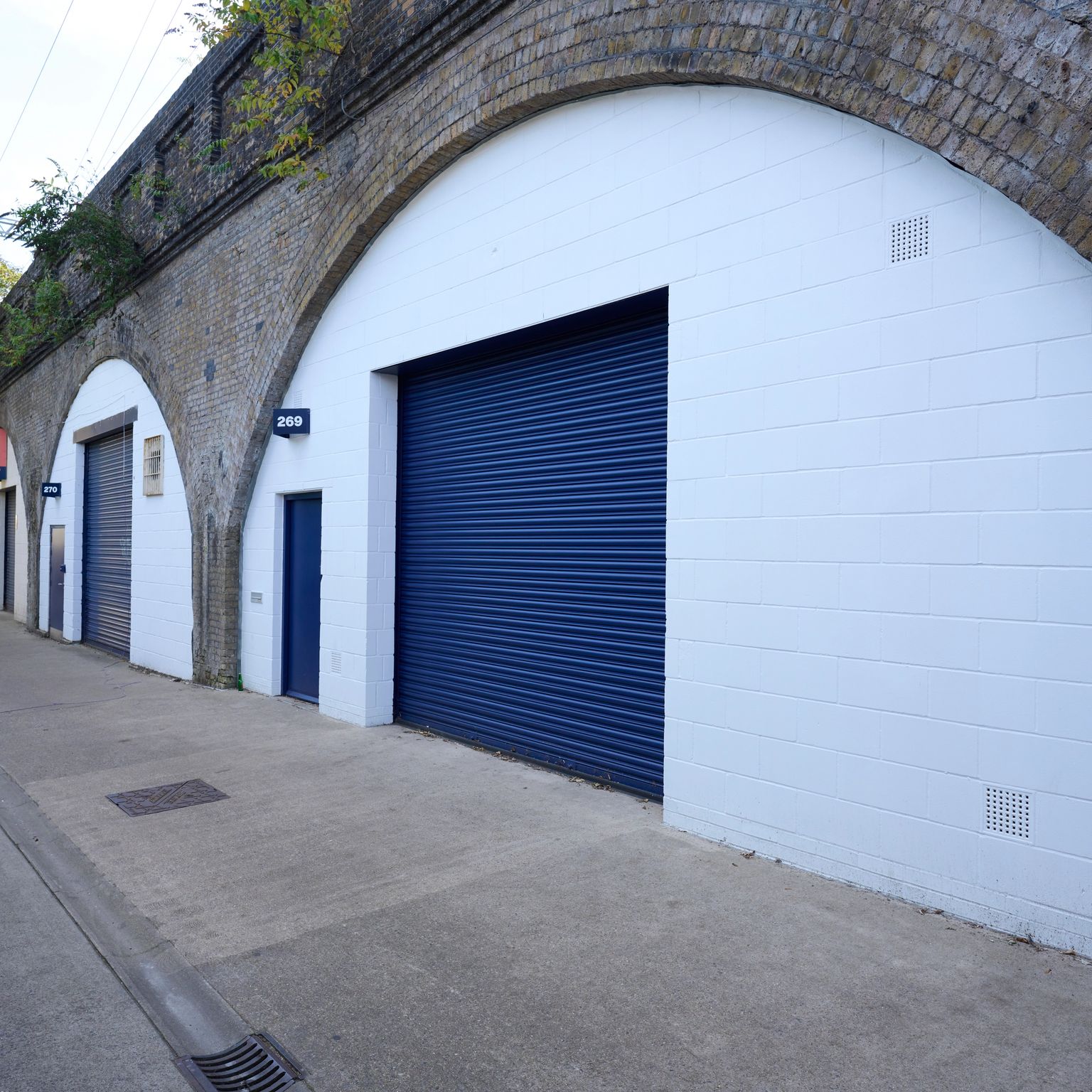 A narrow street alongside brick railway arches converted into white units with dark blue roller shutters.