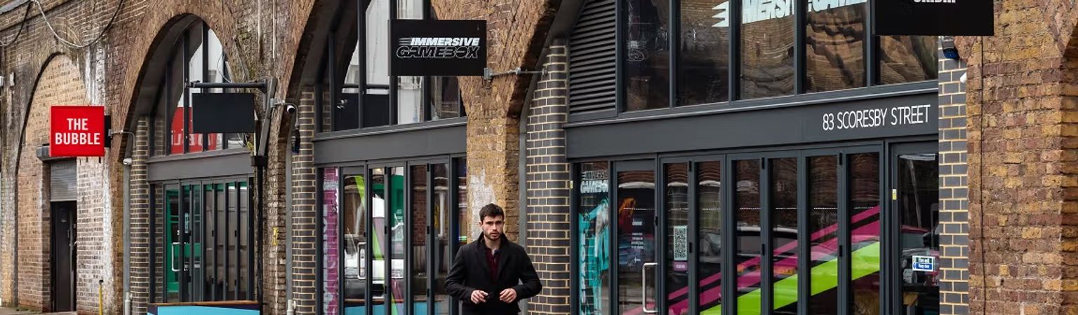 A man walks past storefronts with colorful interiors beneath brick arches, including "The Bubble" and "Immersive Gamebox."