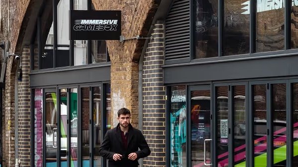 A man walks past storefronts with colorful interiors beneath brick arches, including "The Bubble" and "Immersive Gamebox."