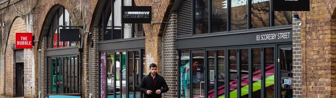 A man walks past storefronts with colorful interiors beneath brick arches, including "The Bubble" and "Immersive Gamebox."