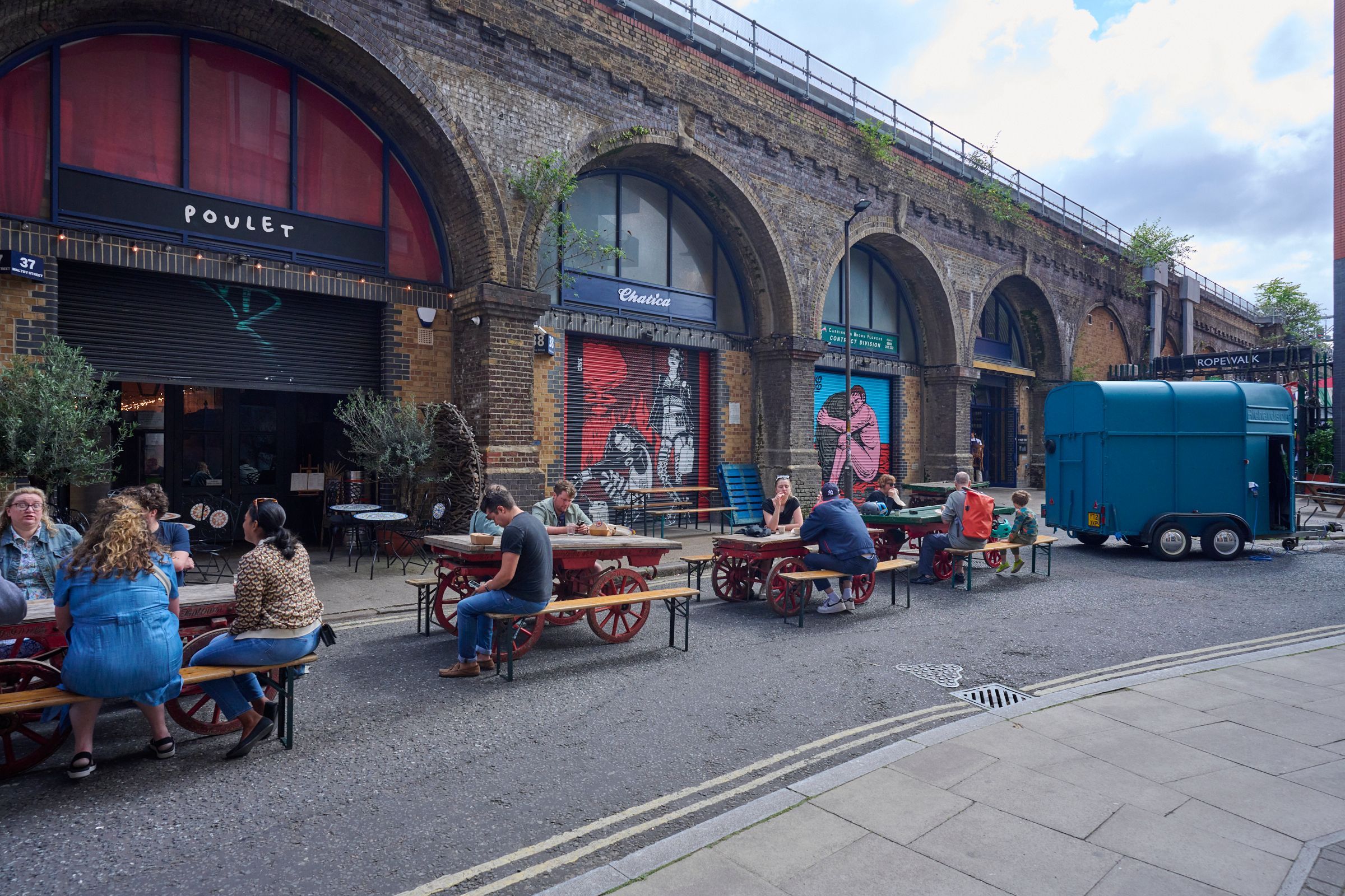 People dine at outdoor tables in front of businesses under brick railway arches, with a blue trailer on the street.