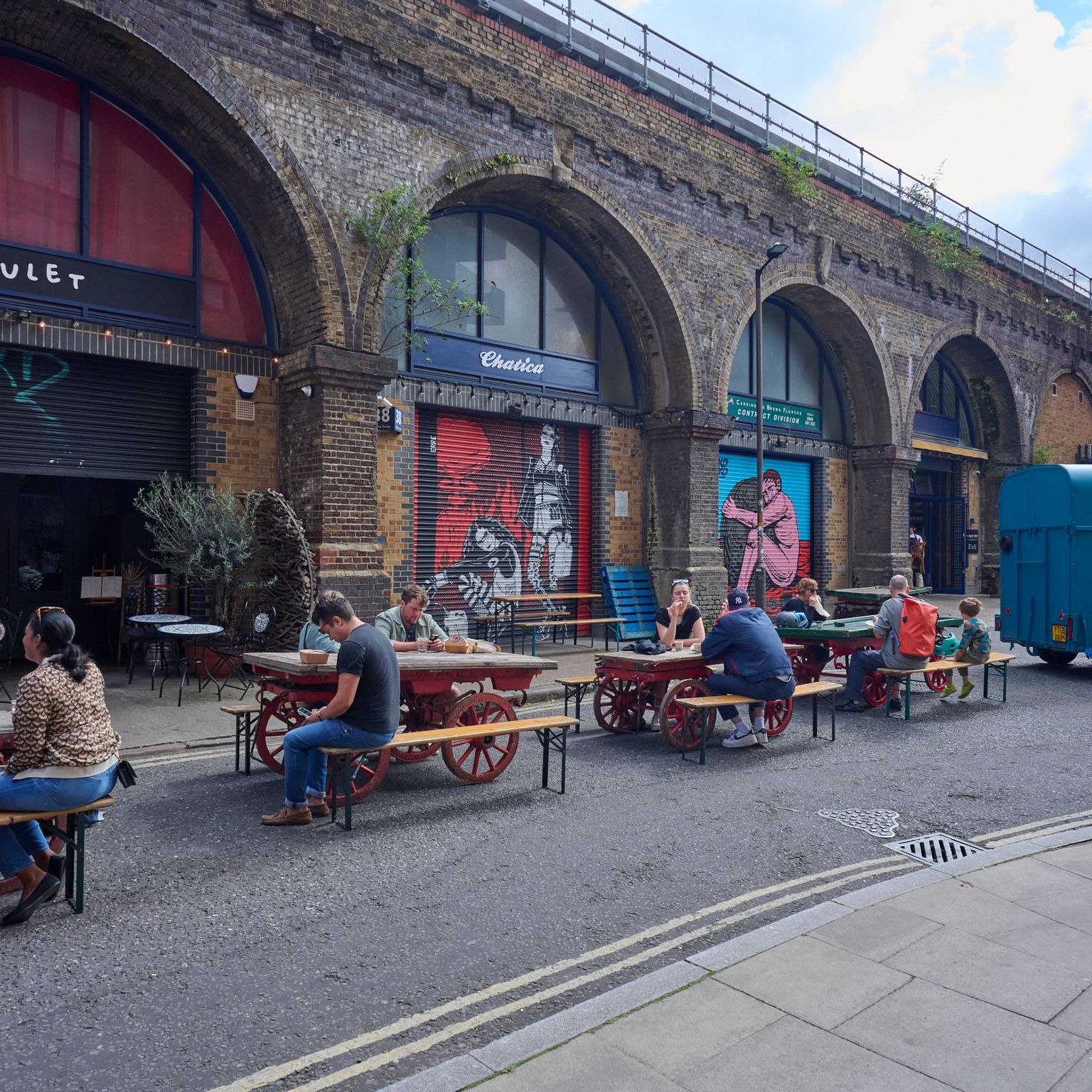 People dine at outdoor tables in front of businesses under brick railway arches, with a blue trailer on the street.