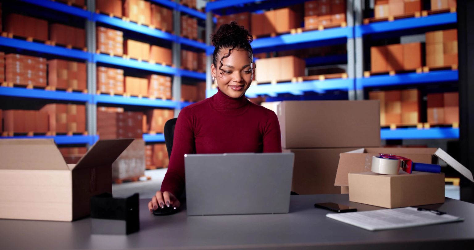A woman works on a laptop at a desk in a warehouse filled with shipping boxes.