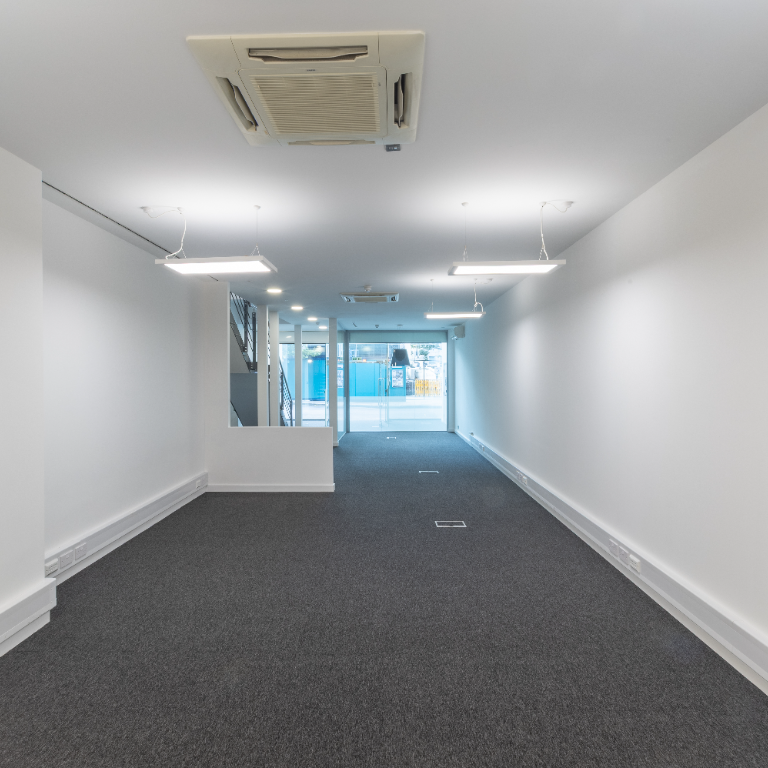 Long, empty commercial unit with gray carpet, white walls, ceiling lights, and a glass storefront entrance.