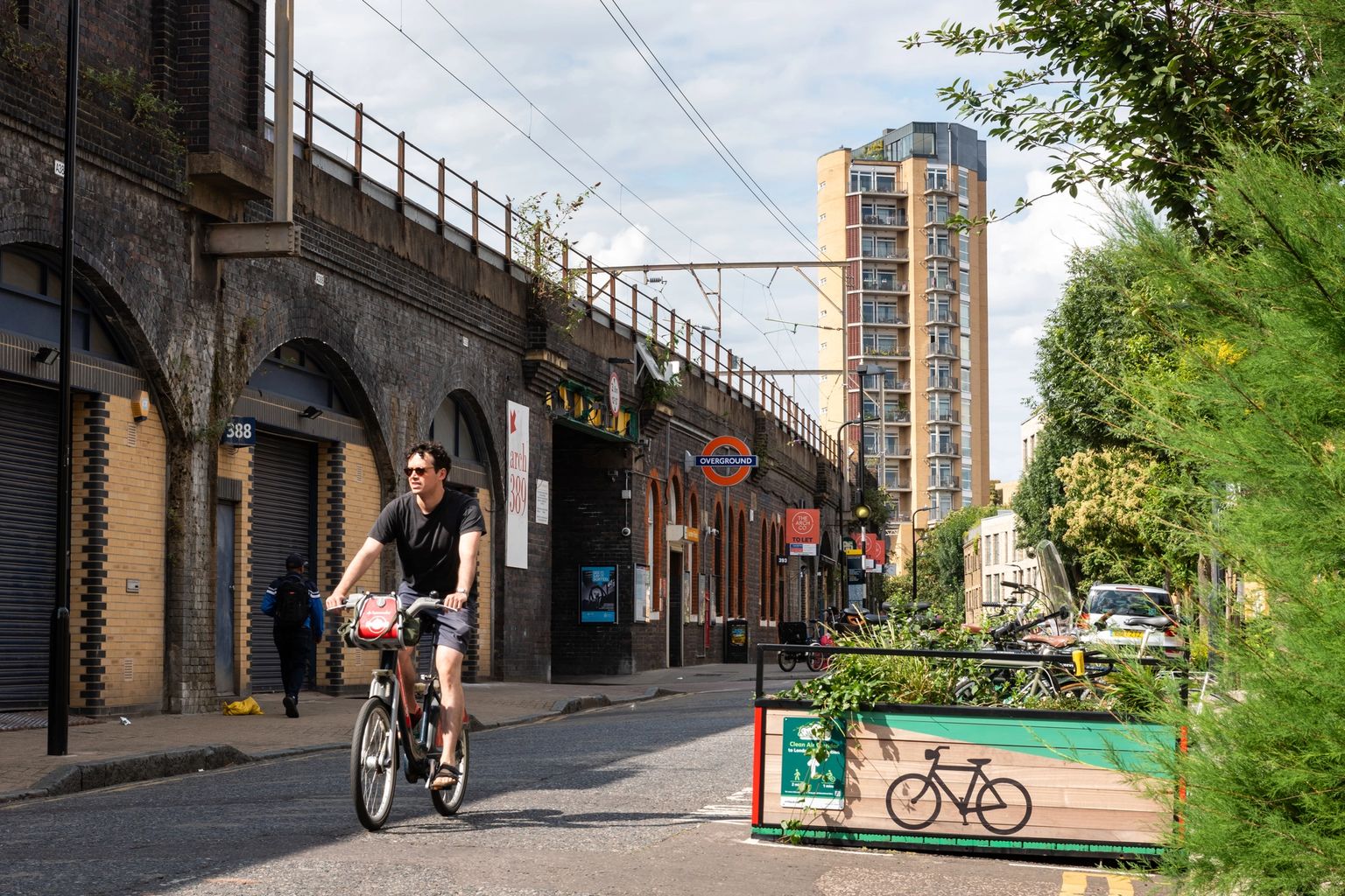A man cycles down an urban street next to a brick railway bridge and a planter with a bike symbol.