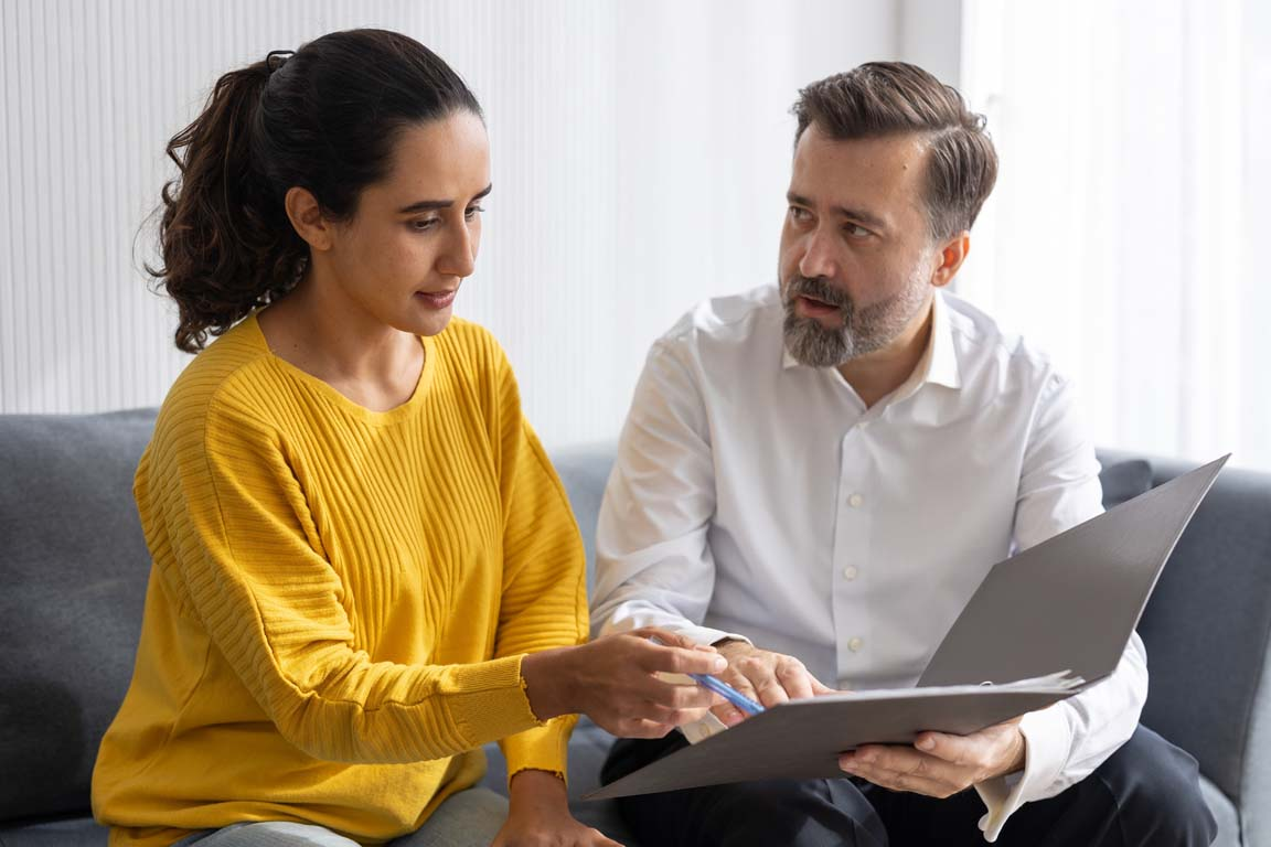 A woman in a yellow sweater points at documents held by a bearded man in a white shirt.