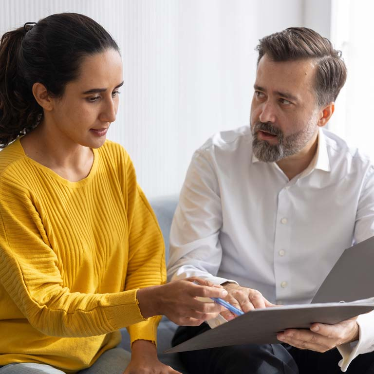 A woman in a yellow sweater points at documents held by a bearded man in a white shirt.