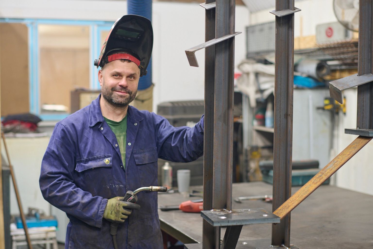 Smiling welder in blue overalls, helmet up, holding a torch next to metalwork.
