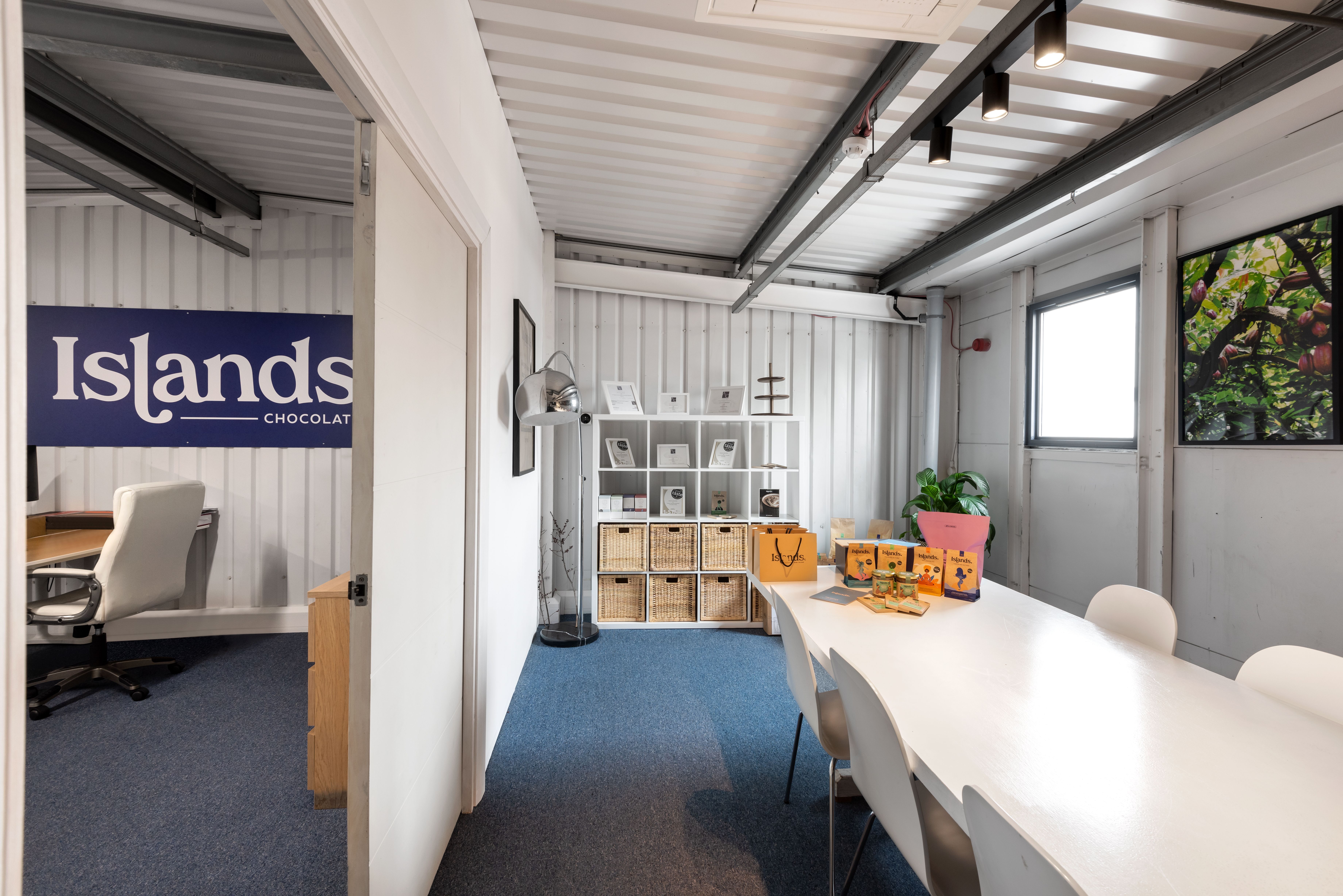An office/meeting room with corrugated white walls, a long table displaying products, and an adjacent room visible through a doorway with an "Islands Chocolat" sign.