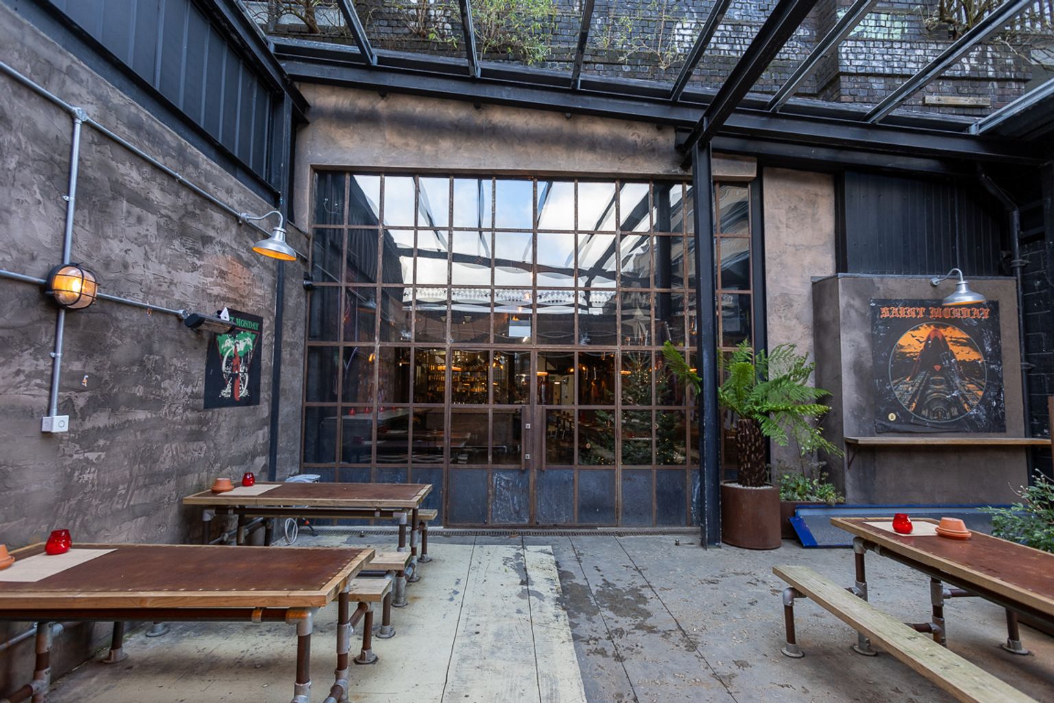 An industrial-style outdoor patio with rustic wooden tables, concrete walls, and large grid-paned glass doors under a metal frame.