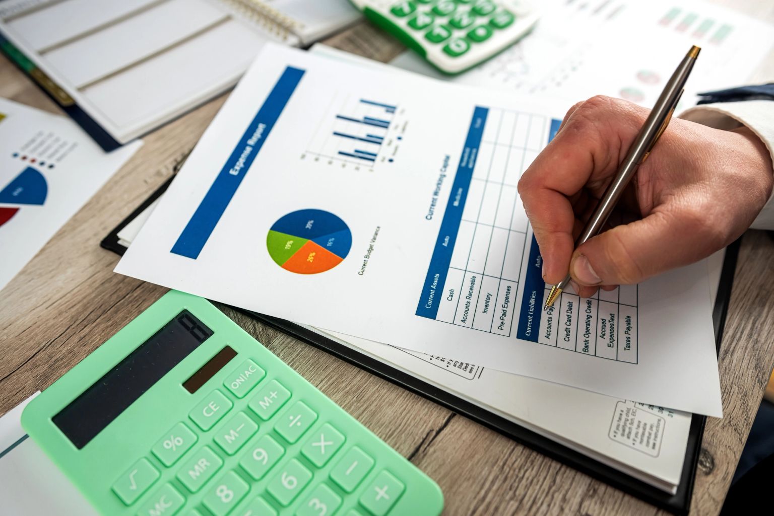 A hand writes on an expense report document with charts and tables, next to a mint green calculator on a wooden desk.