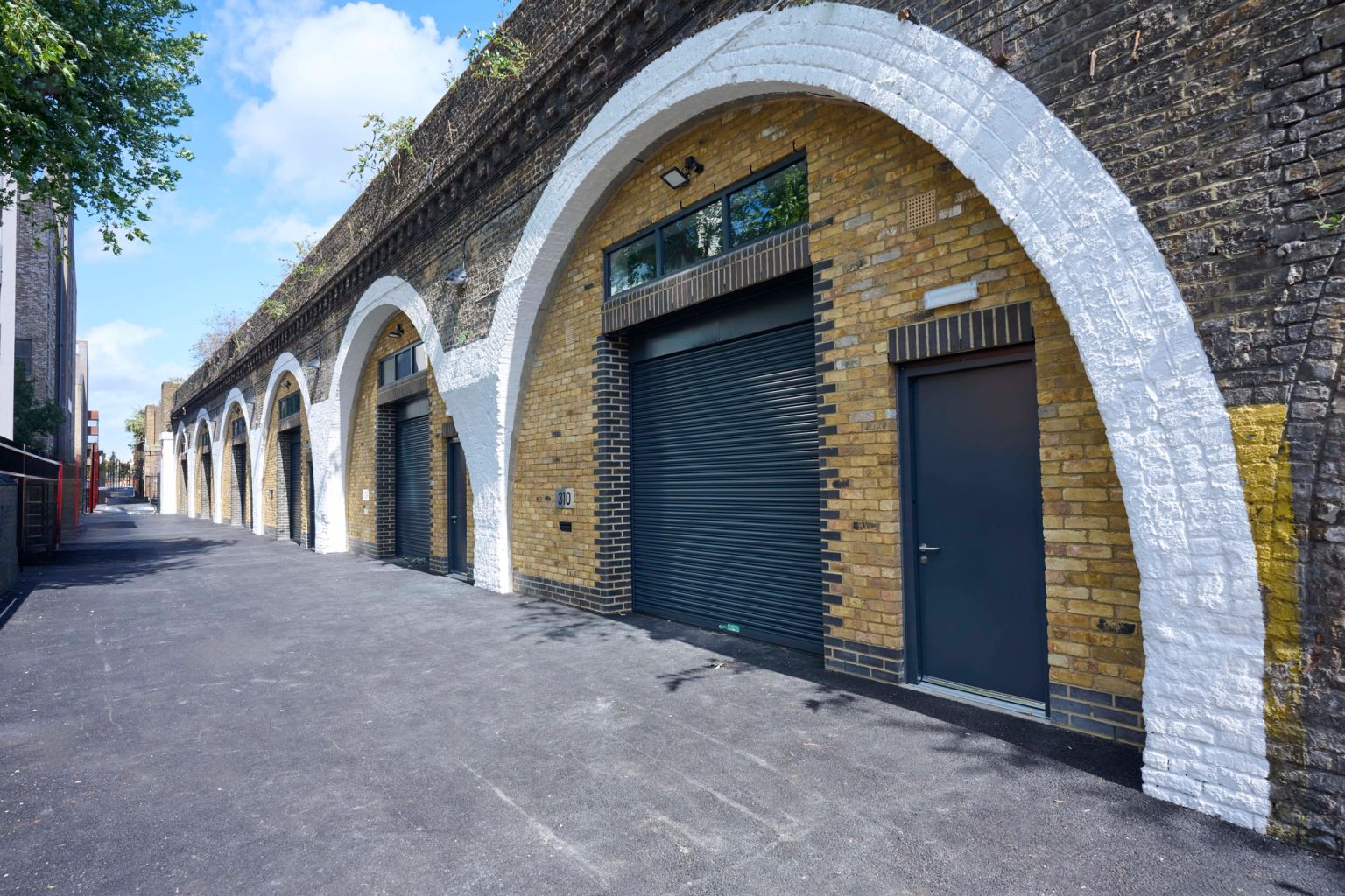 A row of renovated brick railway arches with white trim, featuring dark gray doors and roller shutters, lining a paved walkway.