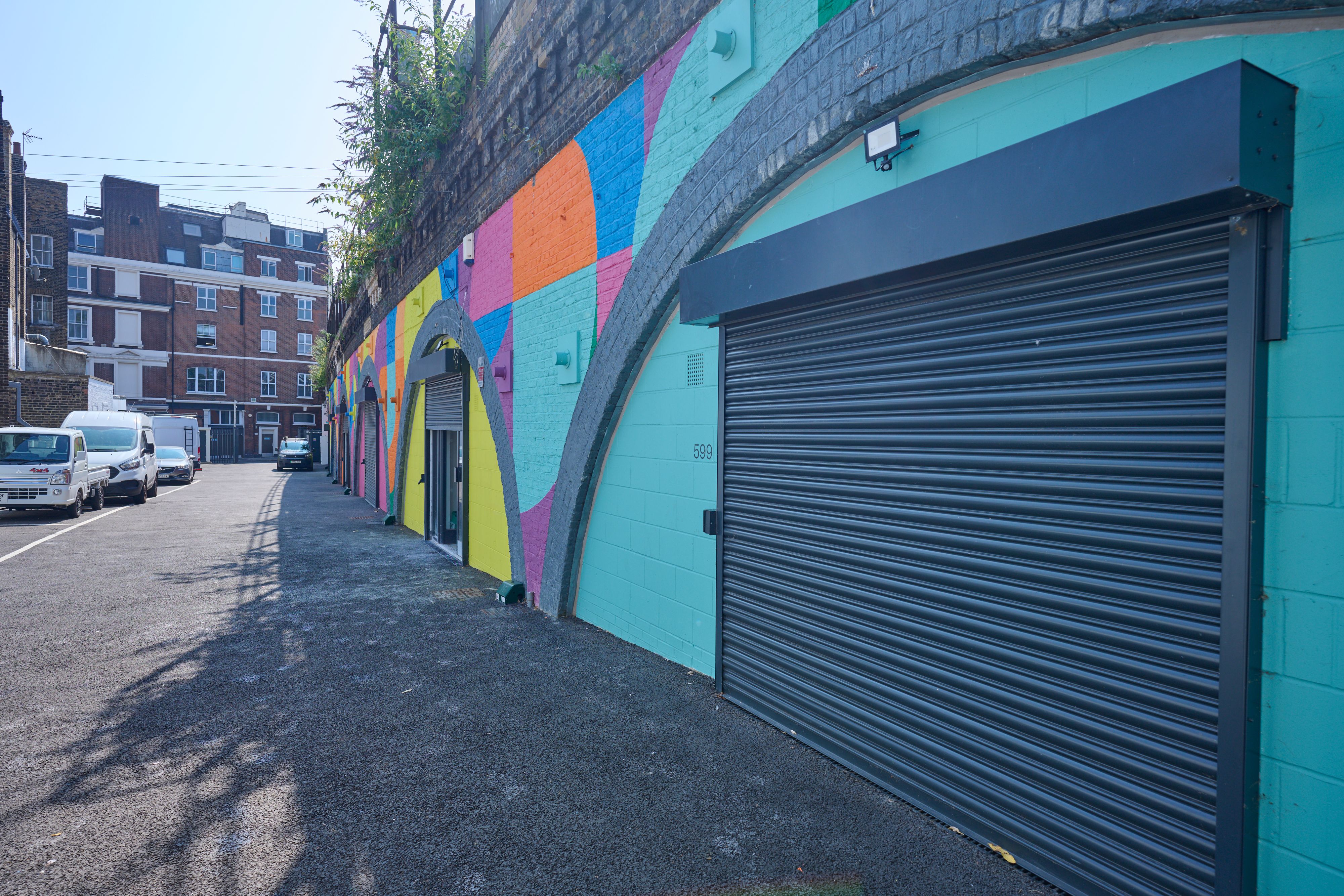 Street alongside a wall with a colorful geometric mural, arched doorways, and a large dark roller shutter.