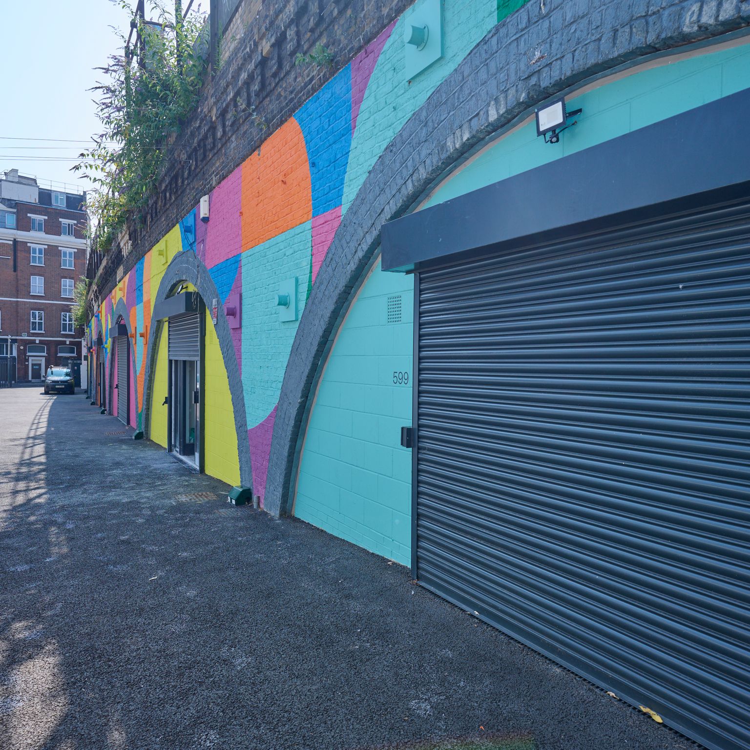Street alongside a wall with a colorful geometric mural, arched doorways, and a large dark roller shutter.