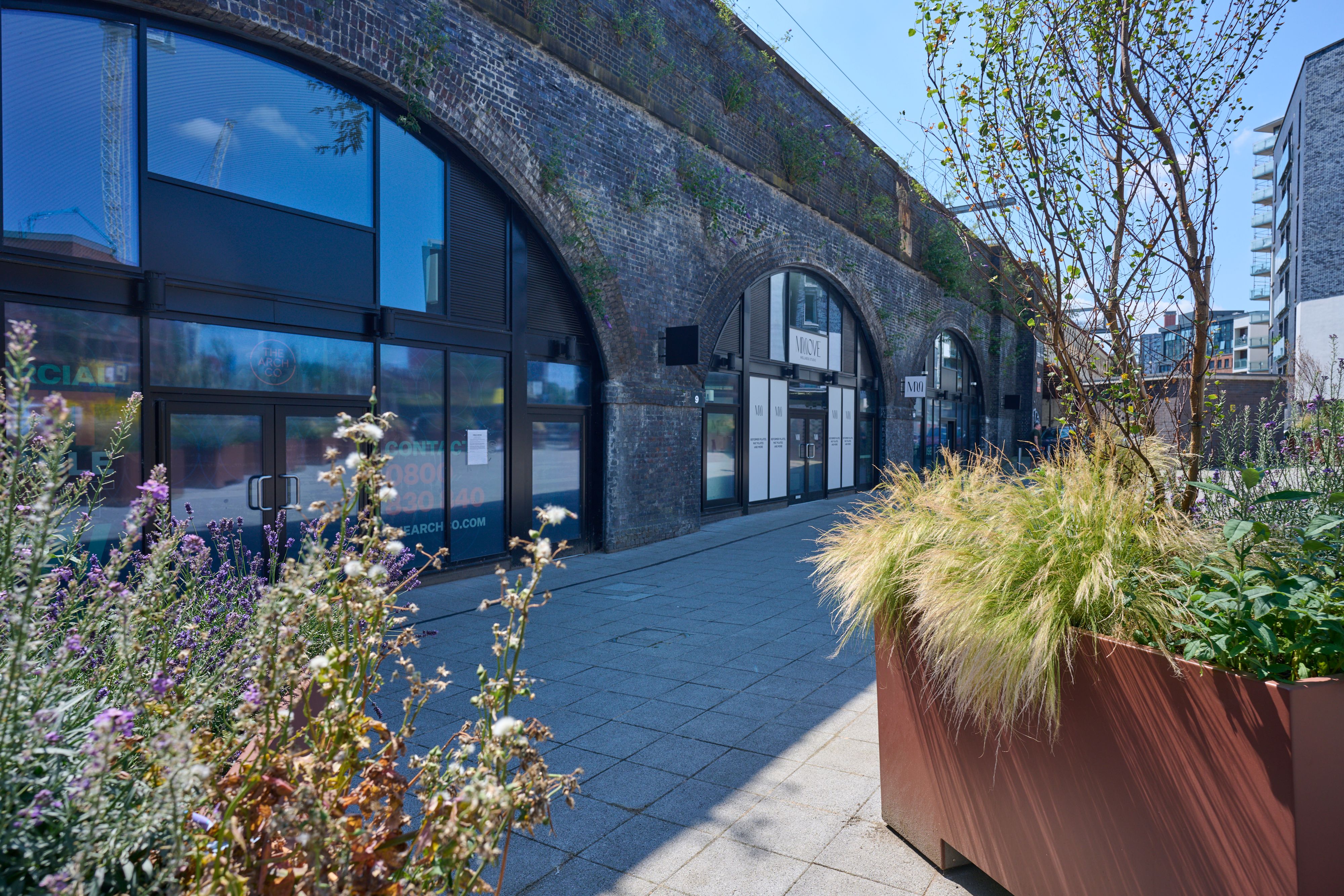 Paved walkway alongside repurposed brick railway arches, with lush planters and modern buildings in the background.