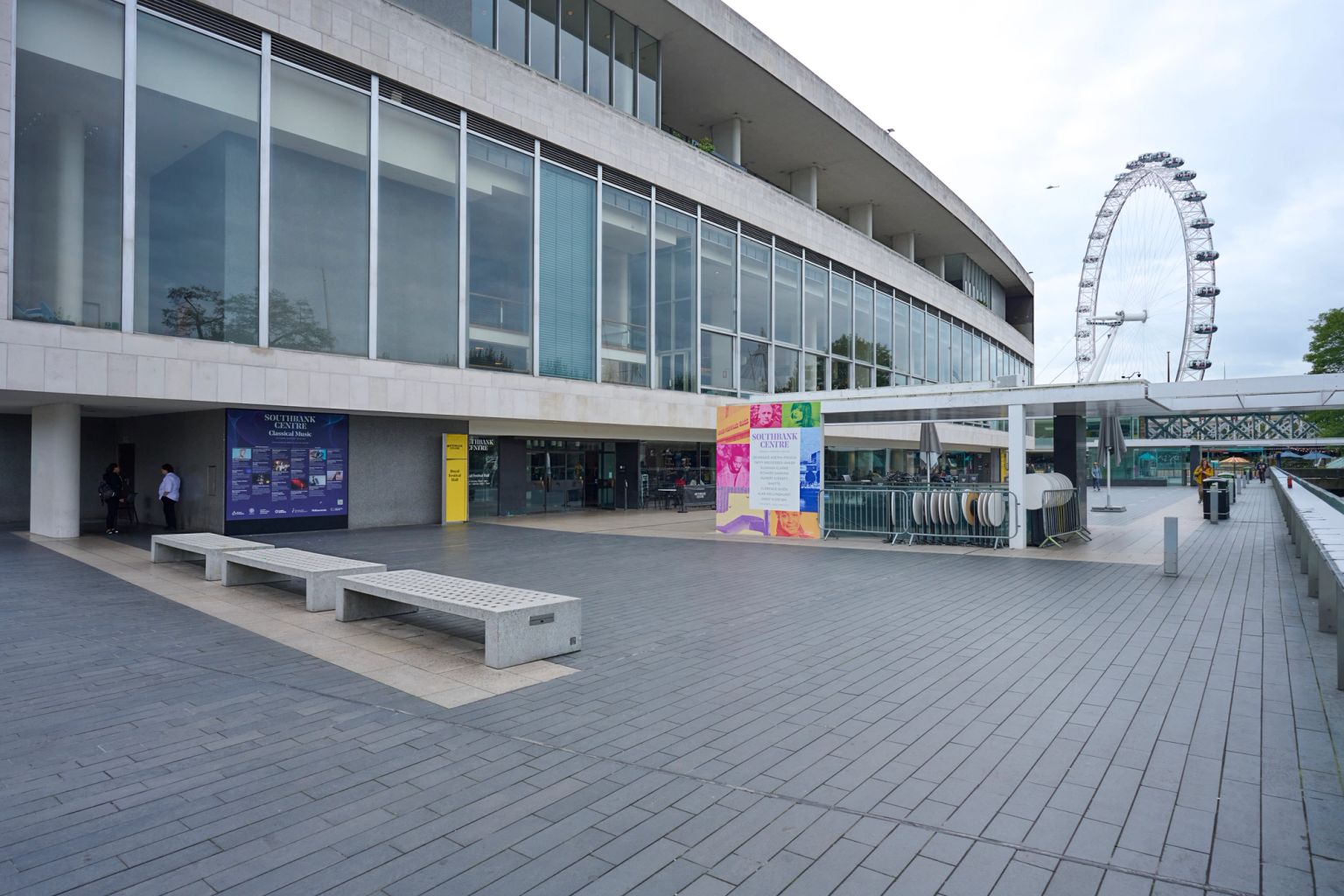 A modern building with large windows, a paved plaza with benches, and the London Eye in the background.