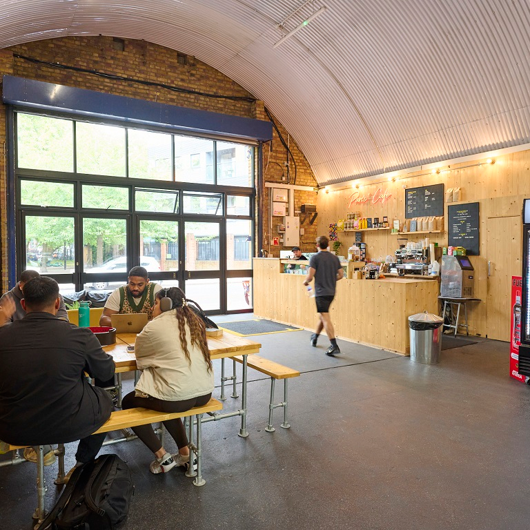 Cafe interior with a vaulted, corrugated metal ceiling, showing customers at a communal table and a service counter.