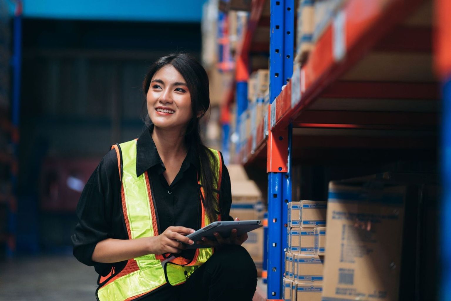 Smiling woman in a safety vest holding a tablet in a warehouse.