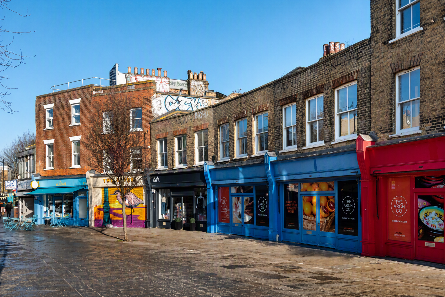 A row of brick buildings with brightly colored shopfronts and a bare tree.