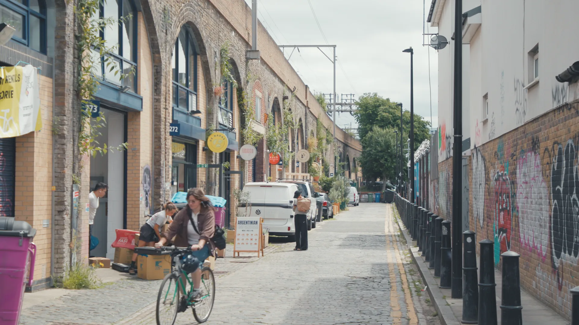 A person cycles down an urban street lined with brick archway shops and a graffiti-covered wall.
