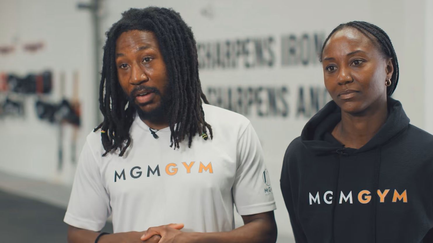 A man with dreadlocks and a woman with braids stand in a gym, both wearing MGMGYM tops.