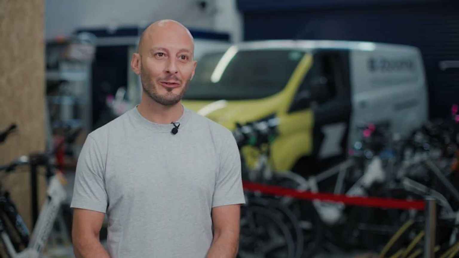 Bald man with a goatee and mic wears a grey t-shirt, standing in a bike shop with a yellow van and bicycles in the background.